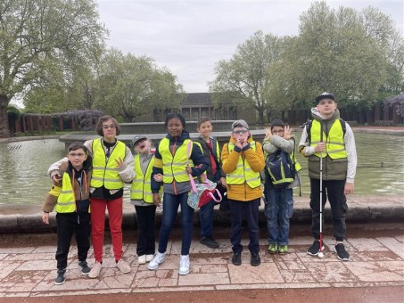 Auf dem Bild ist eine fröhliche Kindergruppe mit gelben Warnwesten zu sehen, die vor einem großen Wasserbecken im Nordpark Düsseldorf stehen. Die Kinder wirken gut gelaunt, einige lachen oder machen lustige Gesten in die Kamera. Es ist auch ein Kind mit einem Langstock zu sehen