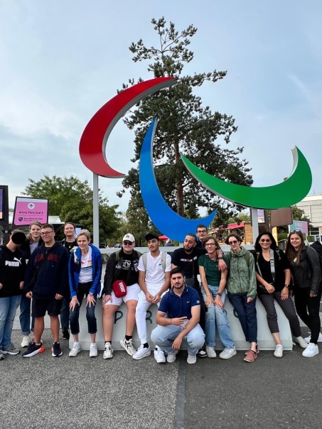 Acht Schüler und 6 Lehrkräfte vor einem Logo der Spiele in Paris.