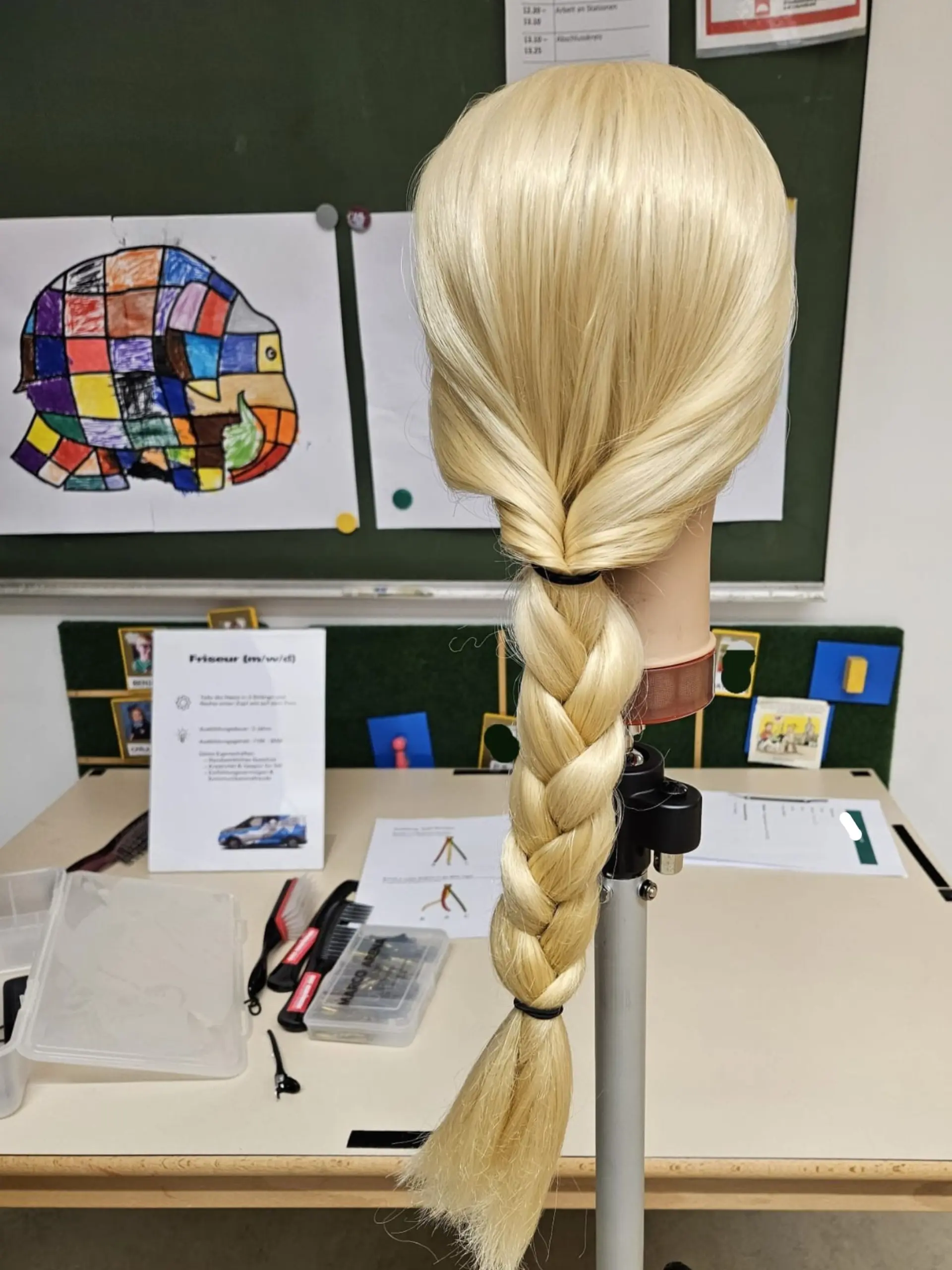 Ein Friseur-Übungskopf mit langen Haaren, die zu einem dicken Zopf geflochten sind. Der Kopf ist auf einem Stativ montiert und steht auf einem Arbeitstisch im Rahmen der Berufsorientierung, auf dem verschiedene Kämme, Bürsten und eine Box mit Haarclips liegen.