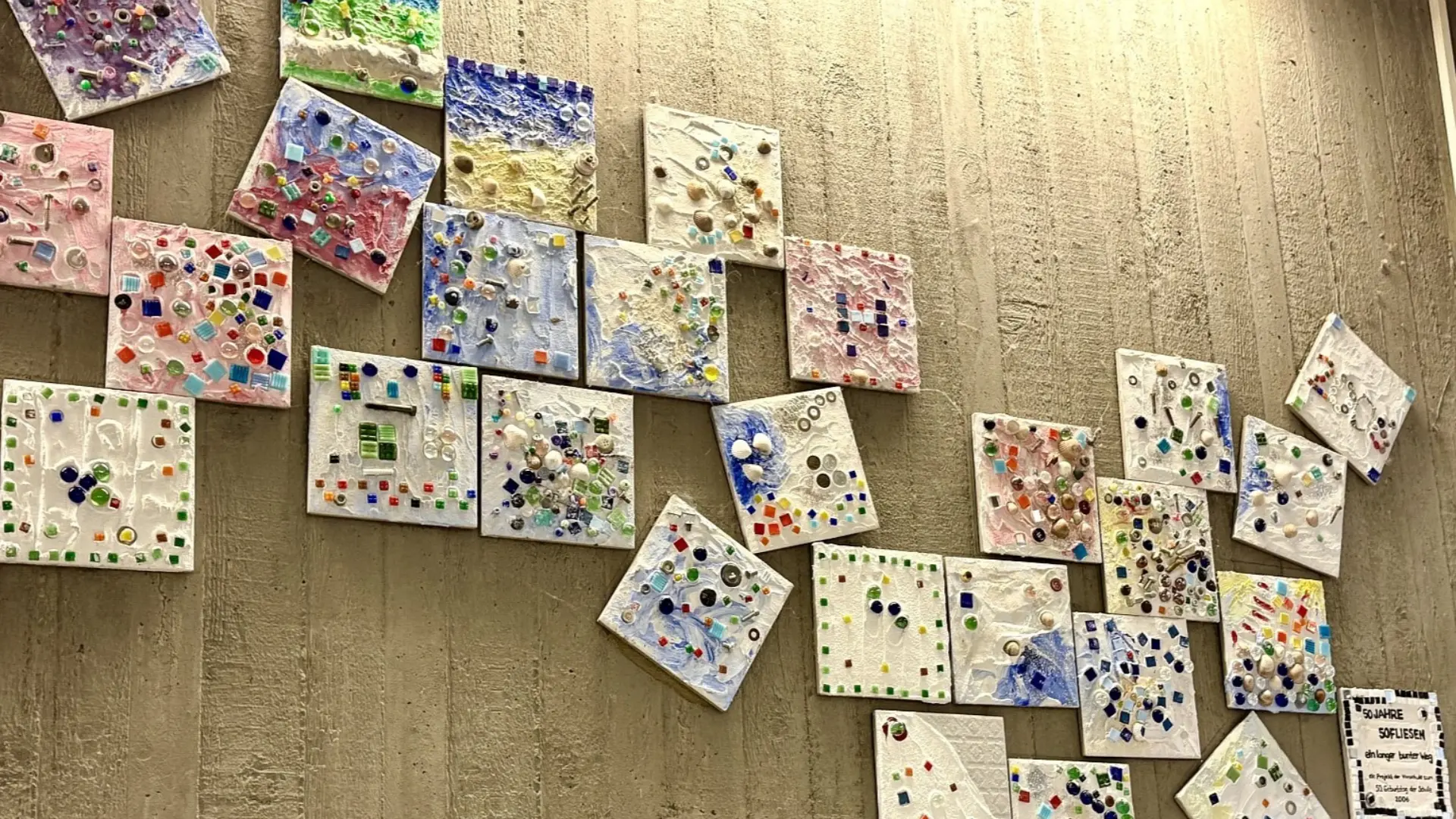 Eine unregelmäßige Anordnung von etwa dreißig quadratischen Mosaikfliesen hängt an einer Betonwand im Flur der Schule. Jede Fliese wurde von Schüler*innen mit bunten Mosaiksteinen, Perlen, Muscheln und Glasstücken individuell gestaltet.