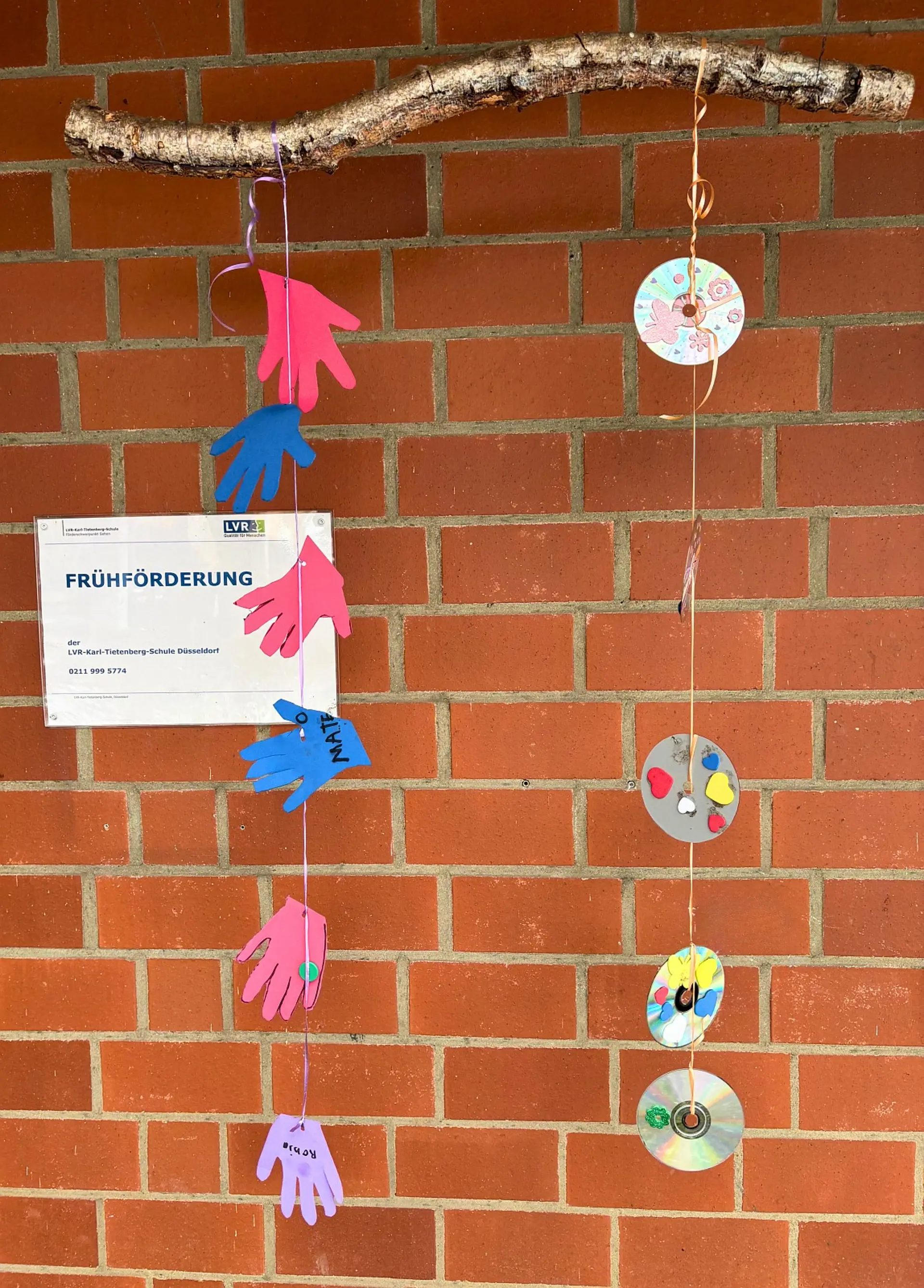 Eine rote Ziegelwand, an der ein waagerechter Ast befestigt ist, von dem zwei dekorative Mobiles herabhängen. Links an der Wand hängt zudem ein Schild mit der Aufschrift Frühförderung der LVR-Karl-Tietenberg-Schule Düsseldorf.