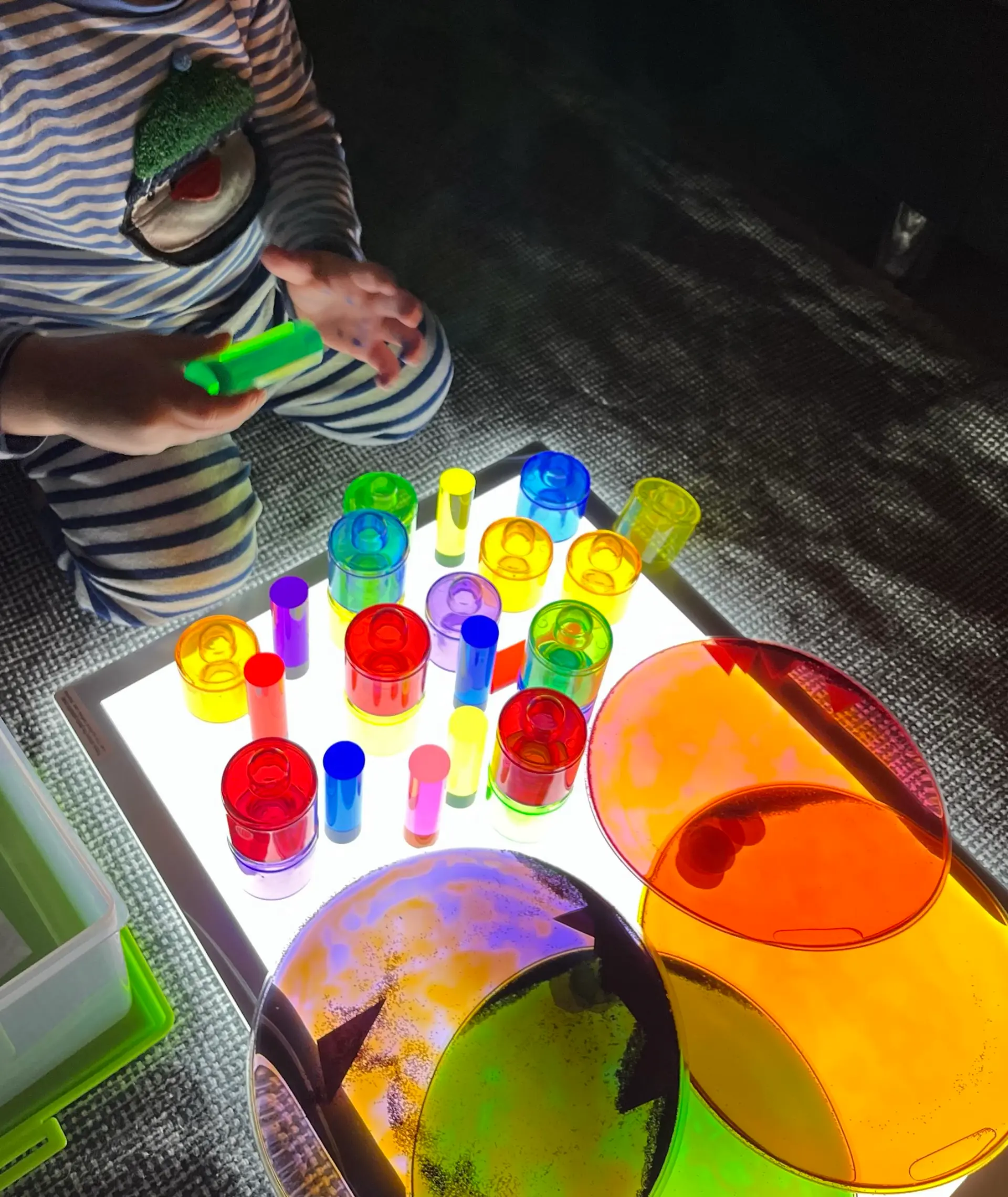 Ein Kind spielt in einem abgedunkelten Raum an einer hellen Leuchtplatte. Auf der Leuchtfläche sind verschiedene transparente, farbenfrohe Spielobjekte verteilt, darunter bunte Zylinder und große, runde Farbscheiben.
