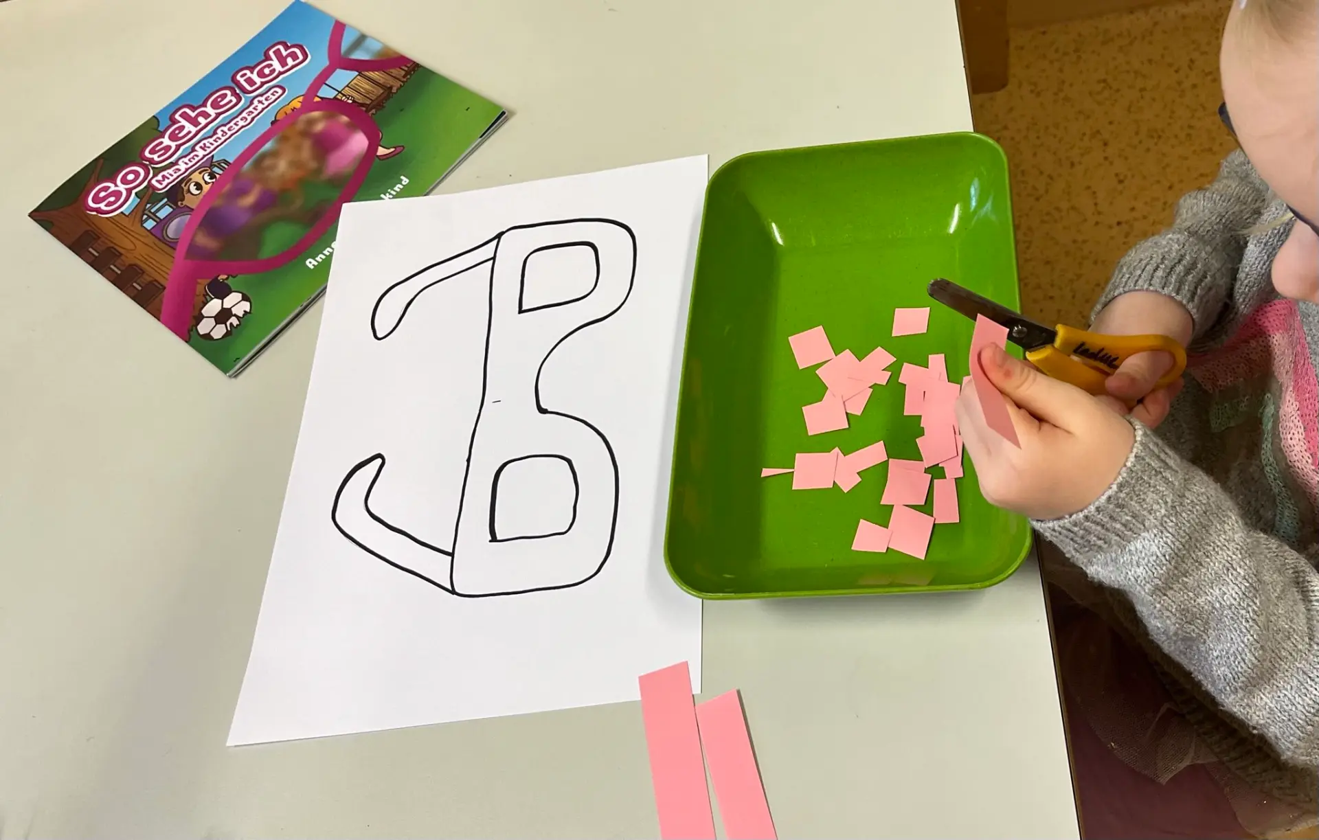 Nahaufnahme eines Kindes bei einer Bastelübung zur Frühförderung. Das Kind nutzt eine Kinderschere, um einen Papierstreifen in kleine Quadrate zu schneiden, die in einer Kunststoffschale gesammelt werden.