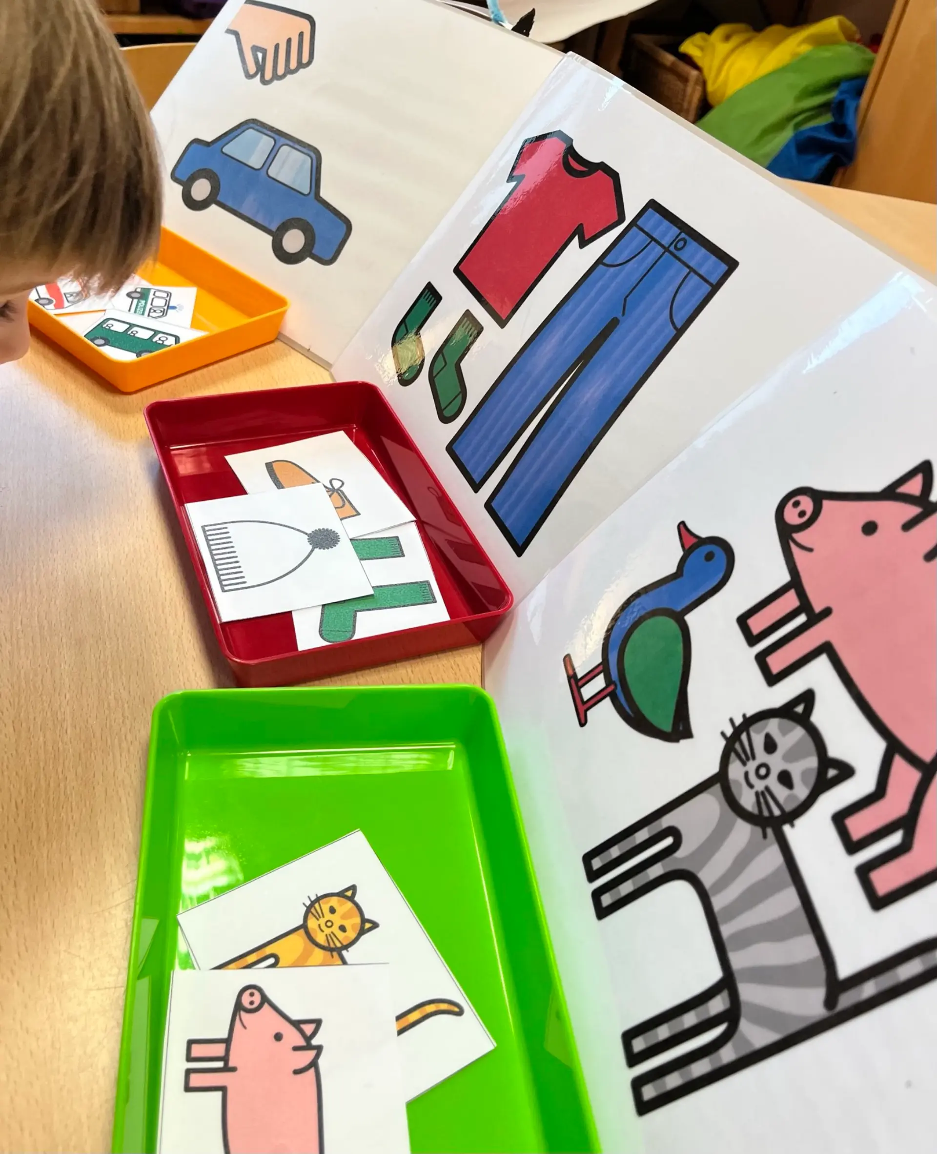 Ein Kind bei einer pädagogischen Sortierübung an einem Holztisch. Vor dem Kind stehen drei farbige Kunststoffschalen, die jeweils einer Kategorie auf einer dahinter stehenden Tafel zugeordnet sind.