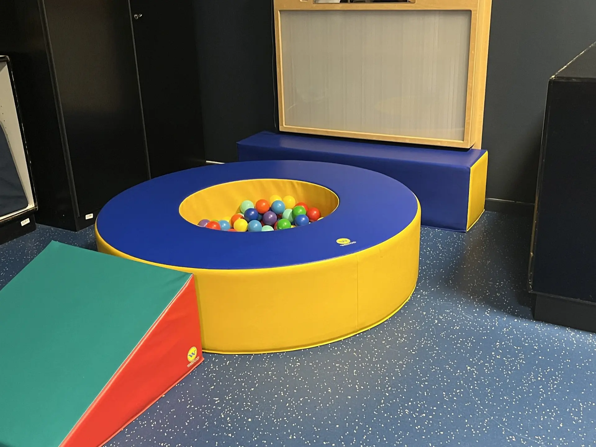 Ein Spiel- und Therapiebereich für die Frühförderung, in dessen Zentrum ein kreisrundes Bällebad steht, das mit bunten Bällen gefüllt ist. Daneben befindet sich eine bunte Schaumstofframpe.