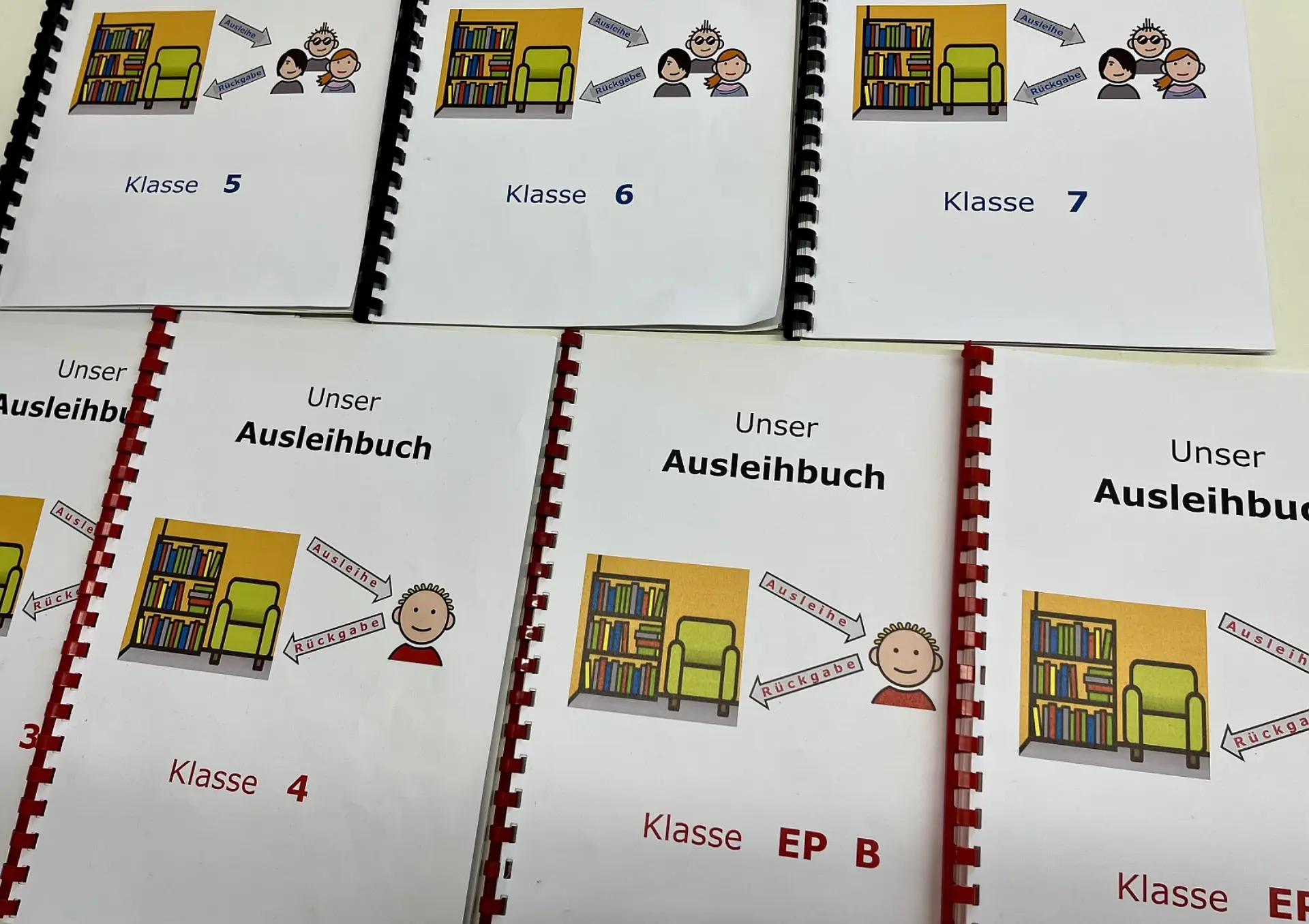 Sieben Ringbuchhefte mit dem Titel Unser Ausleihbuch liegen nebeneinander. Die Hefte sind nach Klassenstufen (z. B. Klasse 4, 5, 6, 7 und EP B) unterteilt. Jedes Deckblatt zeigt eine Metacom-Illustration einer Bibliotheksecke sowie Symbole für Ausleihe und Rückgabe.