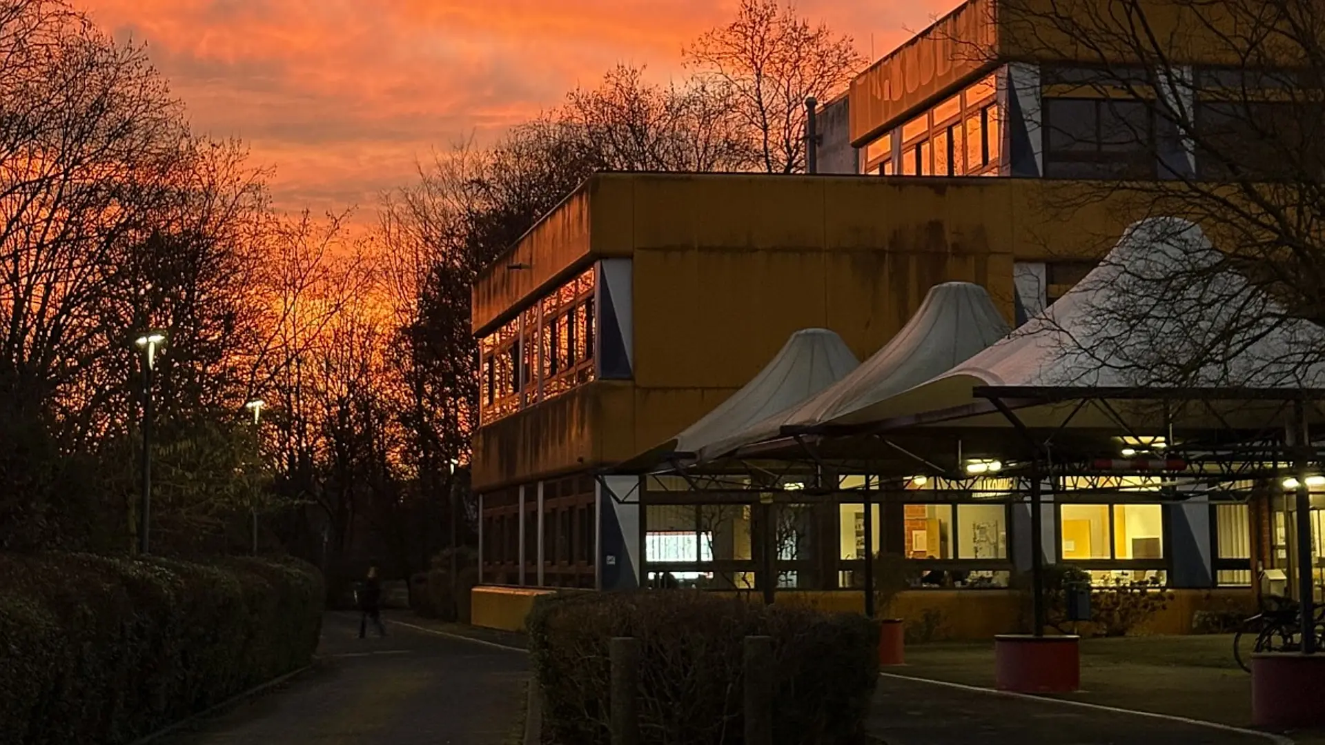 Ein mehrstöckiges Schulgebäude bei einem intensiven, leuchtend orange-roten Sonnenuntergang. Im Vordergrund befindet sich ein gepflasterter Weg.
