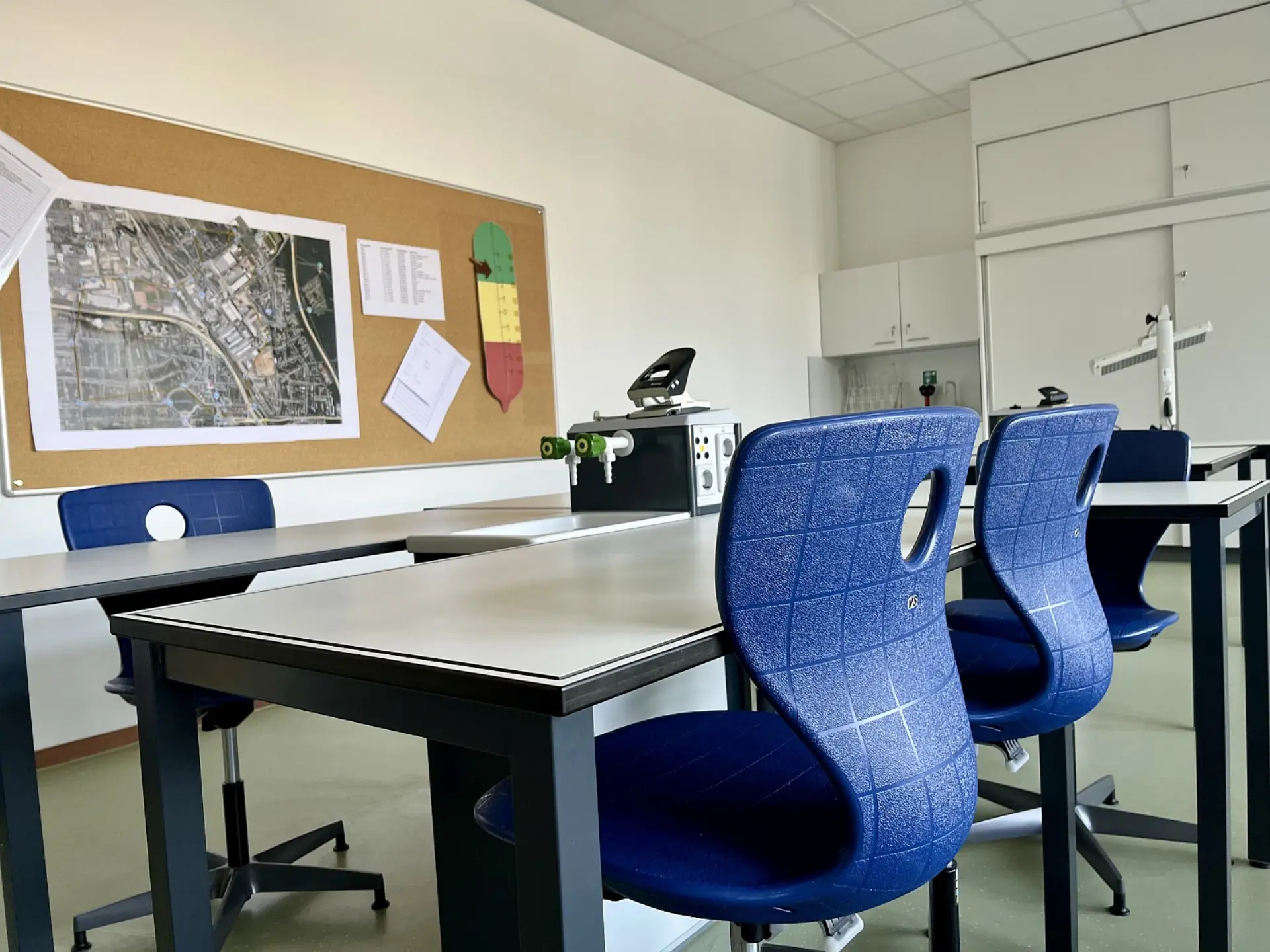 Ein Fachraum für Naturwissenschaften mit ergonomischen Stühlen und Arbeitstischen. An den Tischen befinden sich Versorgungsstationen mit Gasanschlüssen und Steckdosen. An der Wand hängt eine große Kork-Pinnwand mit einer Stadtkarte, Tabellen und einer bunten Lernskala.