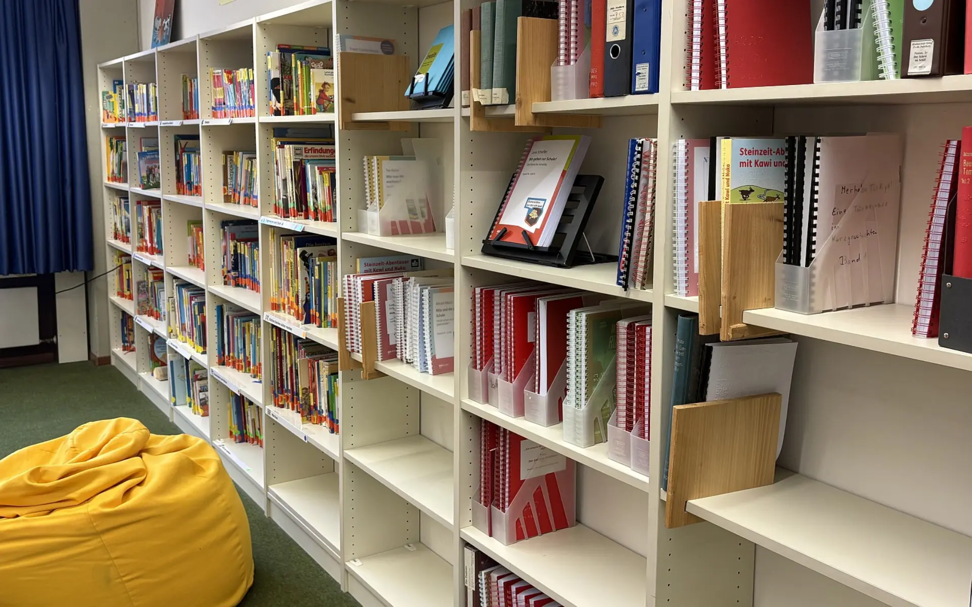 Eine helle, gut sortierte Bücherregal-Wand in der Schülerbücherei. In den Regalfächern stehen zahlreiche Kinder- und Sachbücher. Im Vordergrund links ist ein gemütlicher Sitzsack auf dem Teppichboden zu sehen.