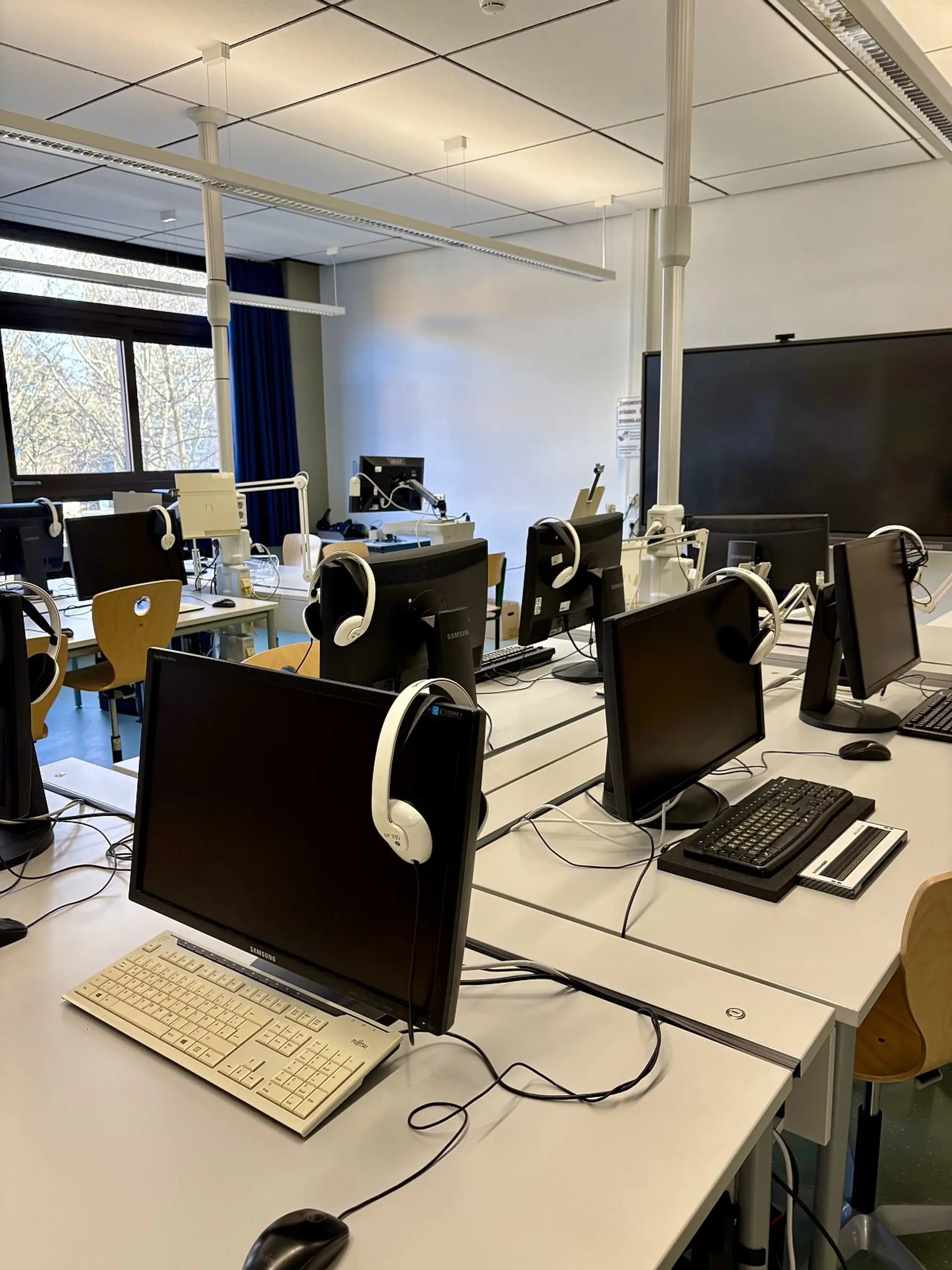 Ein barrierefreier Computerarbeitsplatz in einem PC-Raum. Darin sind mehrere ähnlich eingerichtete Arbeitsstationen mit Monitoren, Tastaturen und Braillezeilen, Halterungen für Dokumente und Kopfhörern zu sehen.