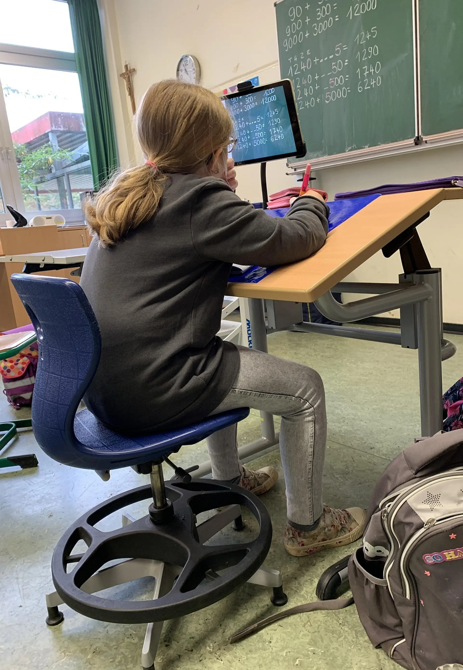 Rückenansicht einer Schülerin, die an einem spezialisierten Schreibtisch im Unterricht arbeitet. Die Tischplatte ist ergonomisch schräg gestellt und mit einer Antirutschmatte versehen. Ein Tablet an einer flexiblen Halterung dient als digitale Sehhilfe und zeigt die Rechenaufgaben der Schultafel in starker Vergrößerung an.