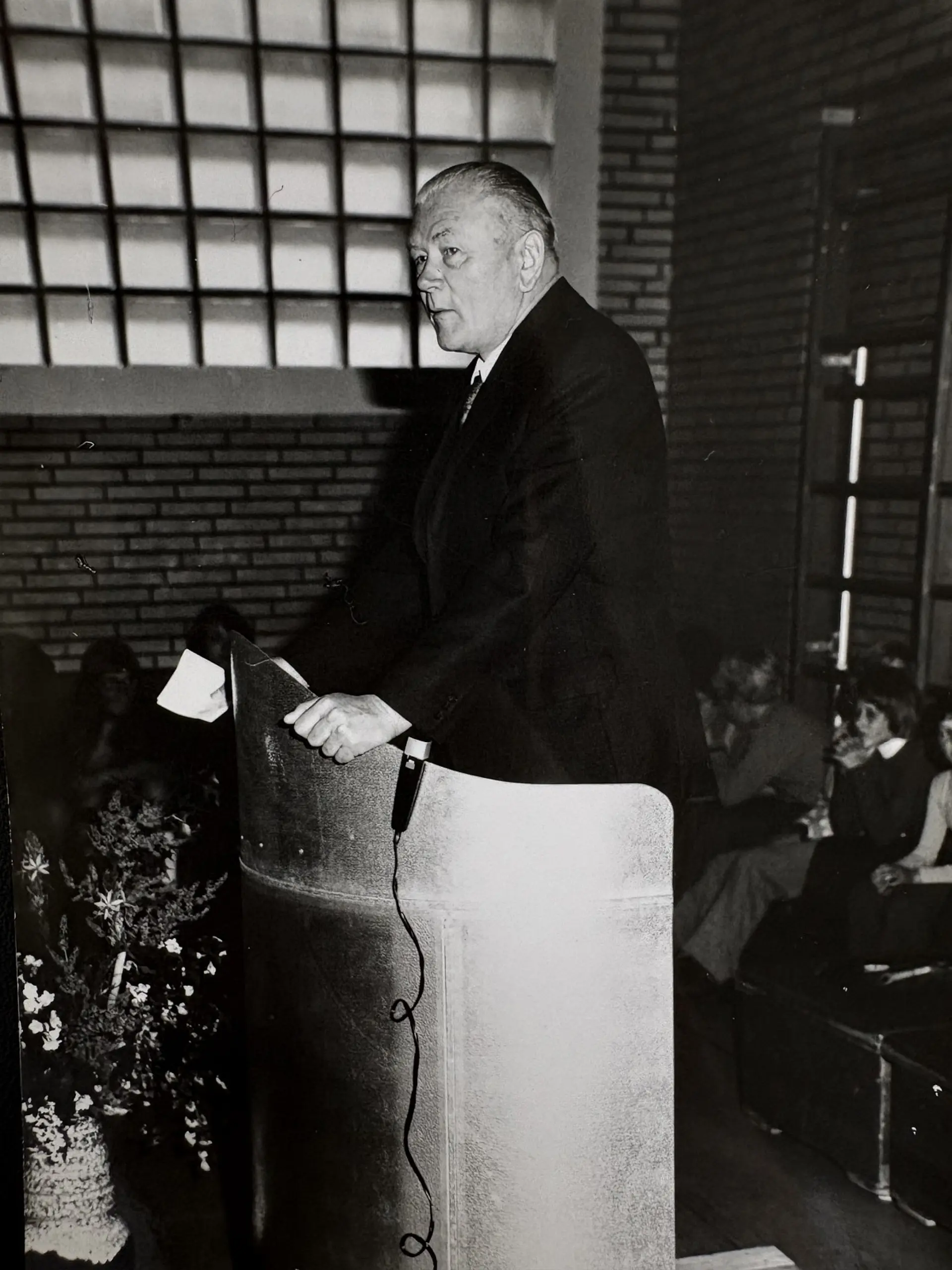 Alte Aufnahme von Karl Tietenberg in Schwarzweiß. Er steht am Sprecherpult in der Turnhalle und hält eine Rede, drum-herum sitzen Lehrkräfte.