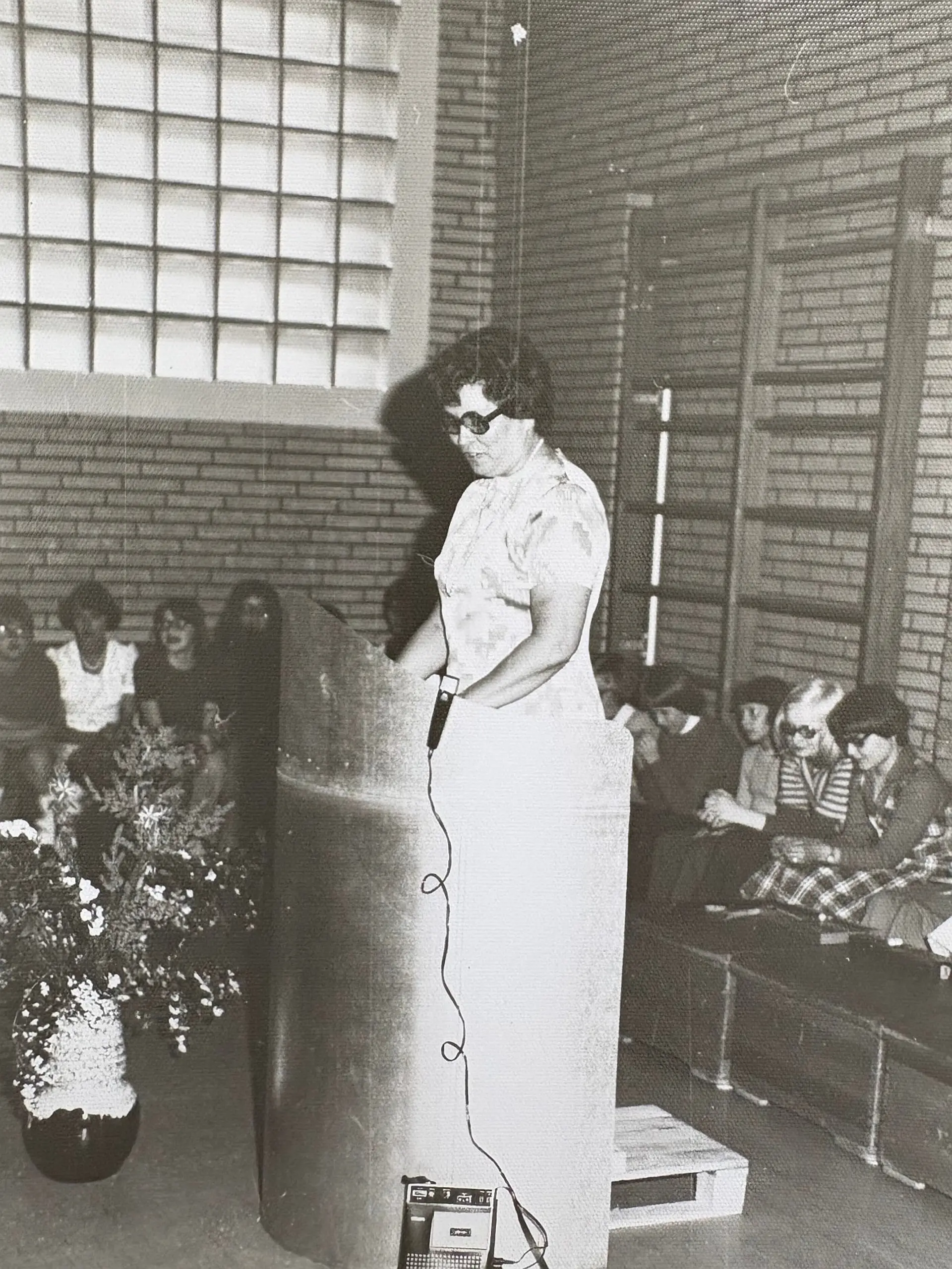Eine alte, schwarzweiße Aufnahmen von der neuen Schulrektorin Maria Neumann. Sie steht am Sprecherpult in der Turnhalle und hält eine Rede. Davor ist ein großer Blumenstrauß, drum-herum sitzen Lehrkräfte.