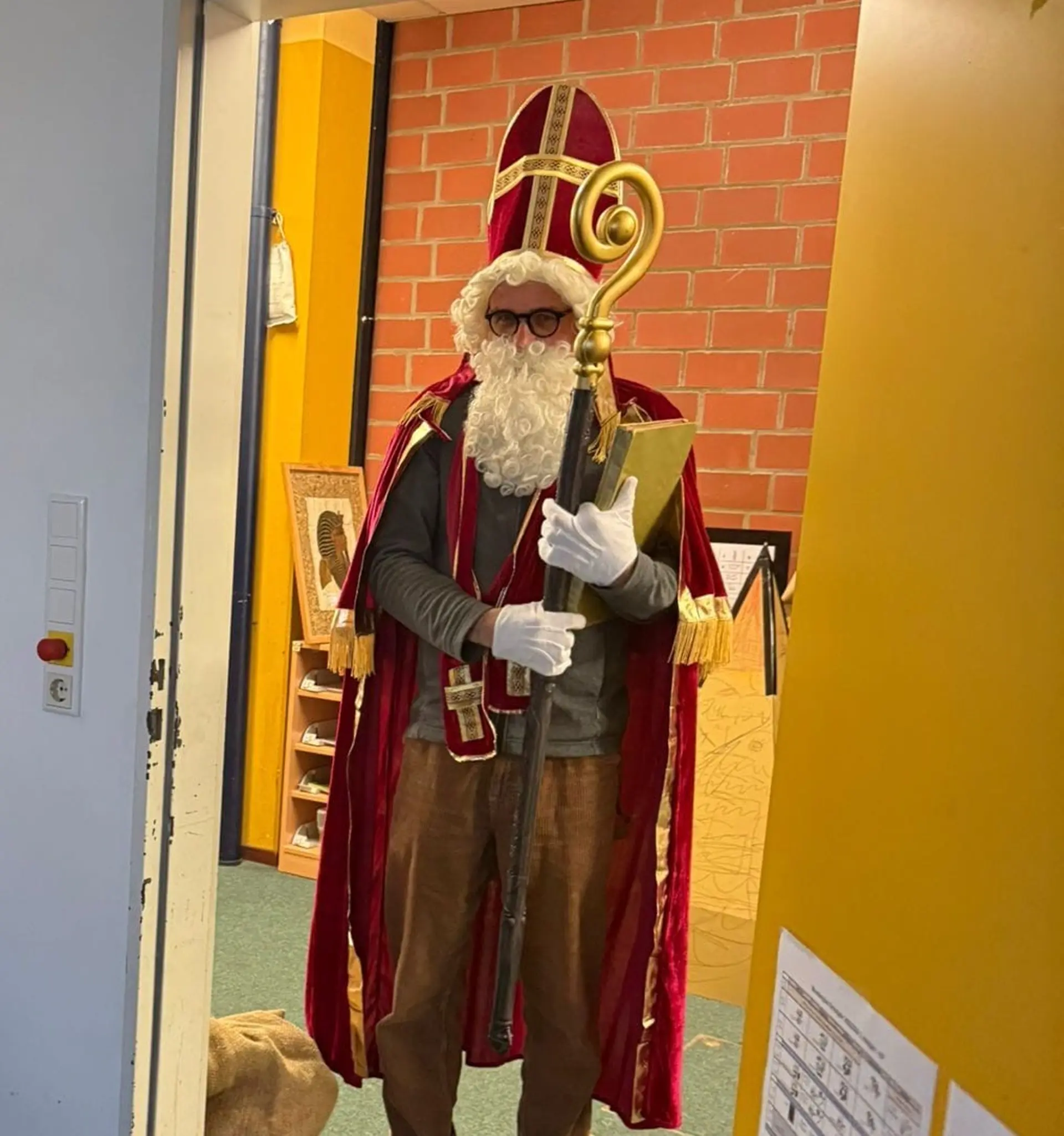 Eine Person im Nikolauskostüm steht in einer Klassenzimmertür. Der Nikolaus trägt eine rote Mitra, einen passenden roten Umhang mit goldenen Verzierungen, einen weißen Rauschebart und weiße Handschuhe. In den Händen hält er einen goldenen Bischofsstab und ein großes goldenes Buch. Neben ihm auf dem Boden steht ein gefüllter Jutesack.