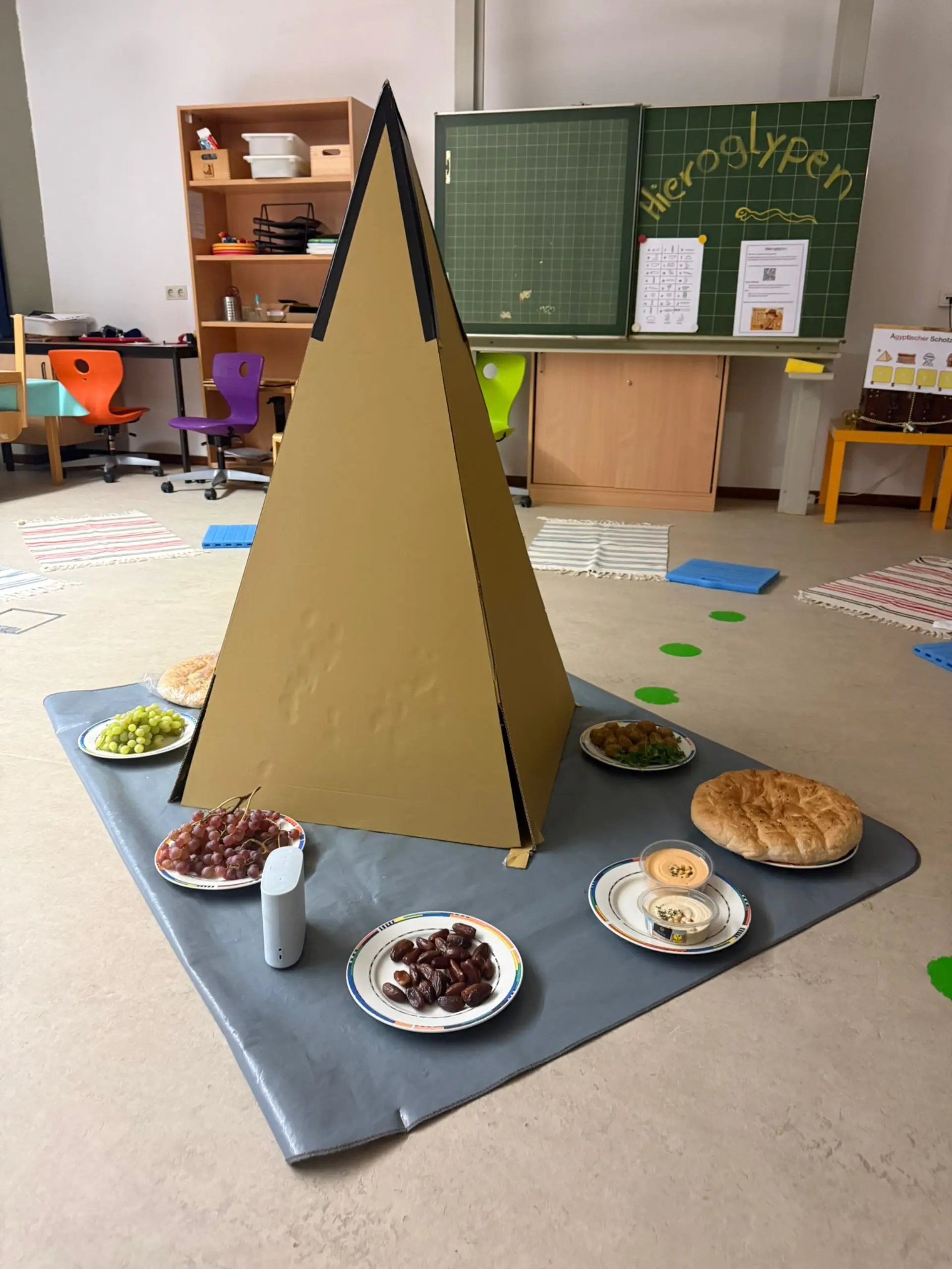 Eine große Pyramide aus Pappe steht im Zentrum eines Klassenzimmers. Um sie herum sind verschiedene Teller mit Speisen wie Weintrauben, Datteln, Fladenbrot, Falafel und Hummus angerichtet. Im Hintergrund ist eine Schultafel zu sehen, auf der das Wort „Hieroglyphen“ steht, sowie bunte Stühle und kleine Teppiche auf dem Boden.