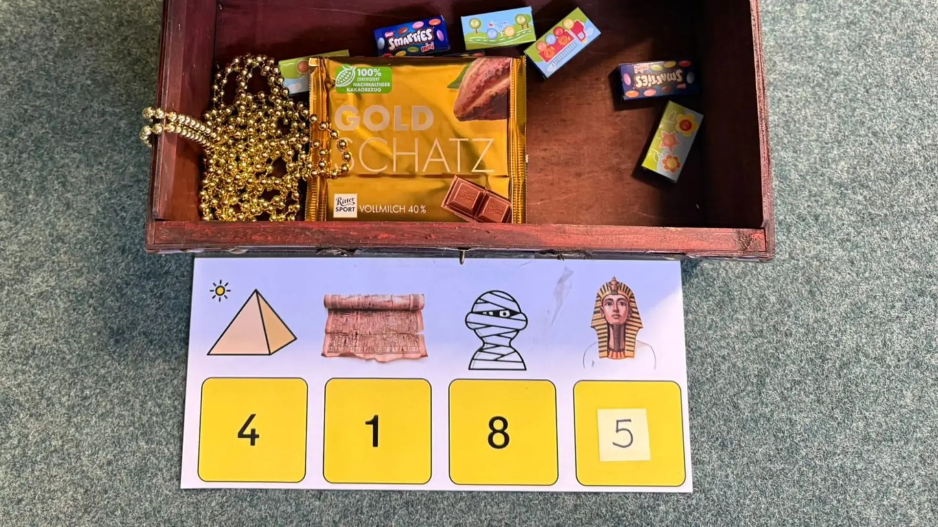 Eine geöffnete Schatztruhe aus Holz mit Lederriemen. Im Inneren liegen eine Perlenkette, eine Tafel Schokolade mit der Aufschrift „Goldschatz“ sowie mehrere kleine Süßigkeiten-Schachteln. Vor der Truhe liegt eine Karte mit vier ägyptischen Symbolen, denen jeweils eine Ziffer als Code zugeordnet ist.