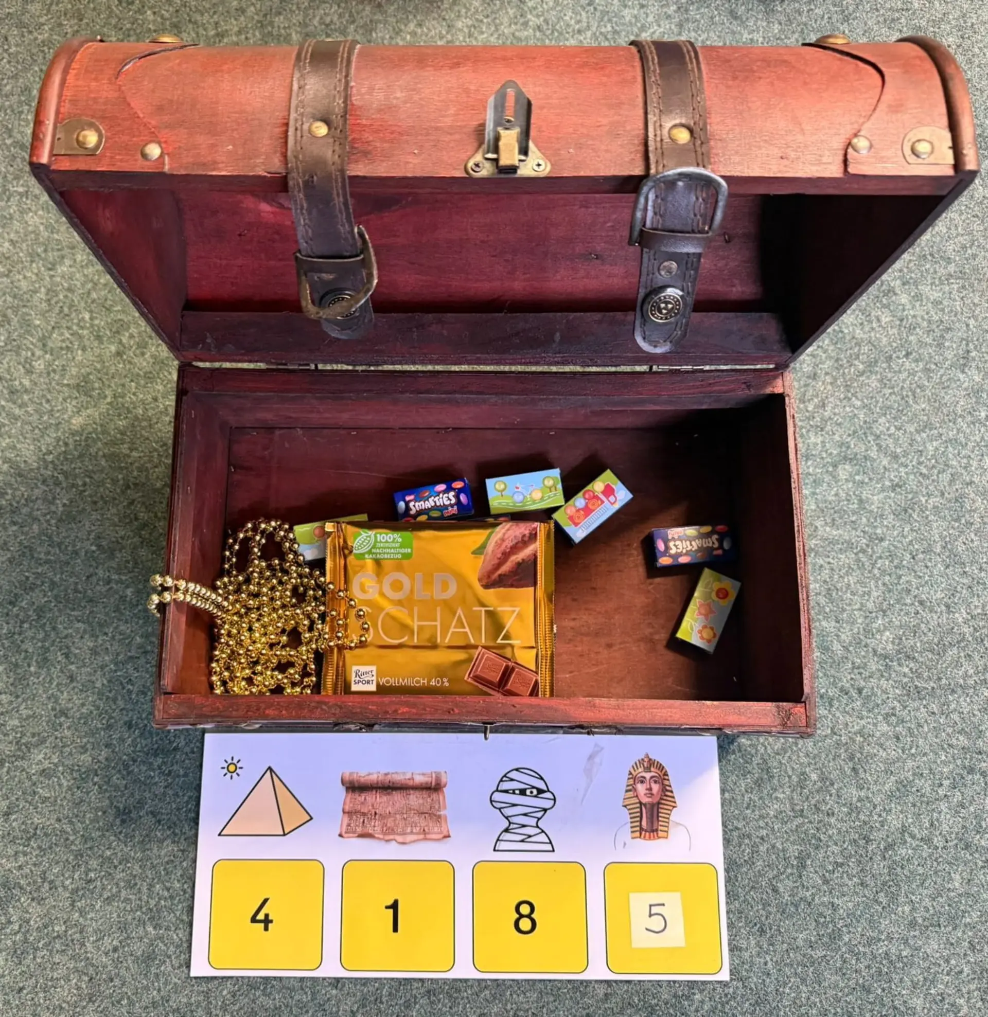 Eine geöffnete Schatztruhe aus Holz mit Lederriemen. Im Inneren liegen eine Perlenkette, eine Tafel Schokolade mit der Aufschrift „Goldschatz“ sowie mehrere kleine Süßigkeiten-Schachteln. Vor der Truhe liegt eine Karte mit vier ägyptischen Symbolen, denen jeweils eine Ziffer als Code zugeordnet ist.