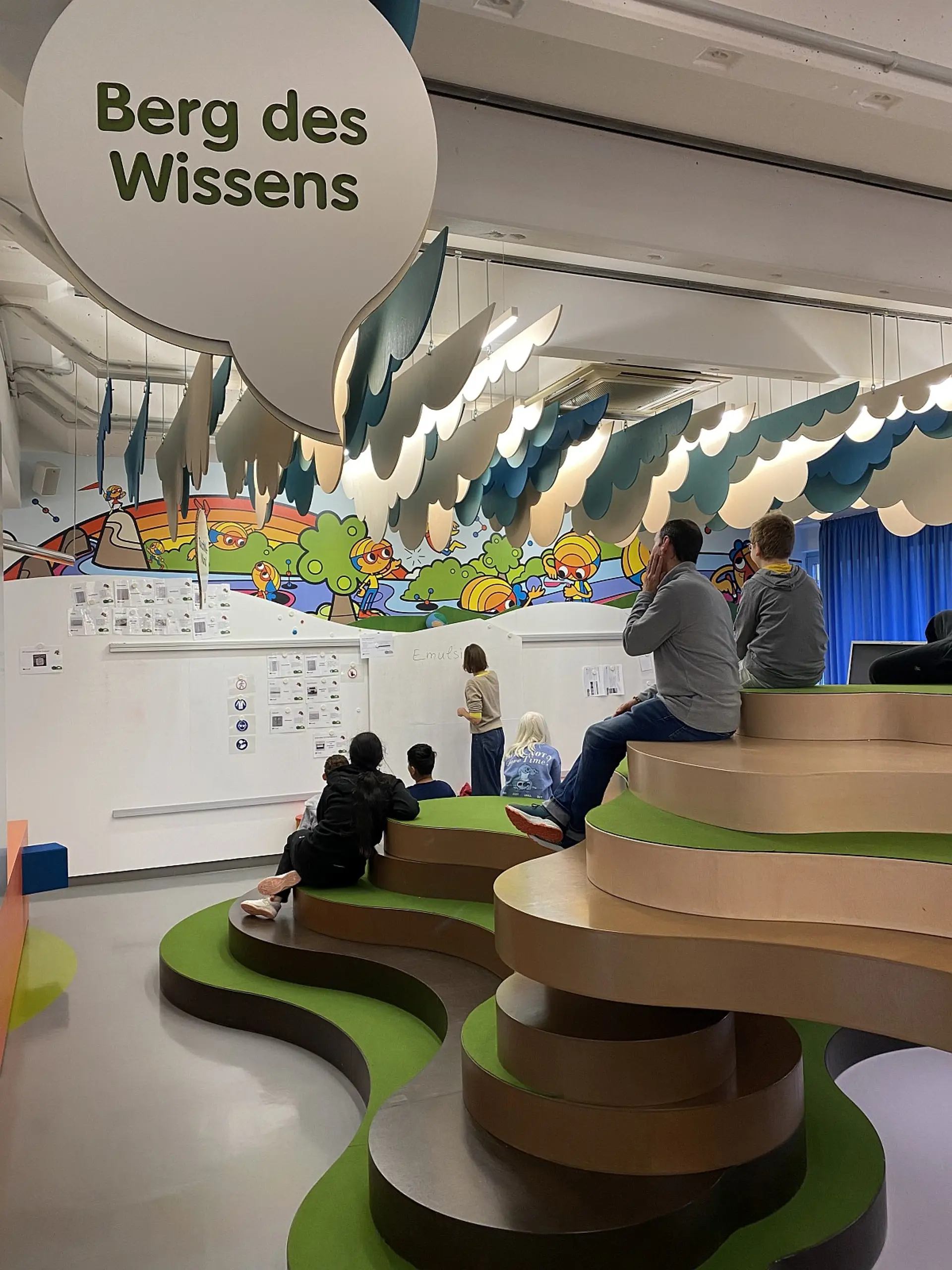 Ein Lernbereich namens „Berg des Wissens“ mit einer wellenförmigen, terrassenartigen Sitzlandschaft aus Holz und grünen Flächen. Mehrere Schüler*innen sitzen auf den Stufen und blicken auf eine Wand mit Schautafeln und bunten Illustrationen. Davor steht eine erwachsene Person und schreibt an der Tafel.