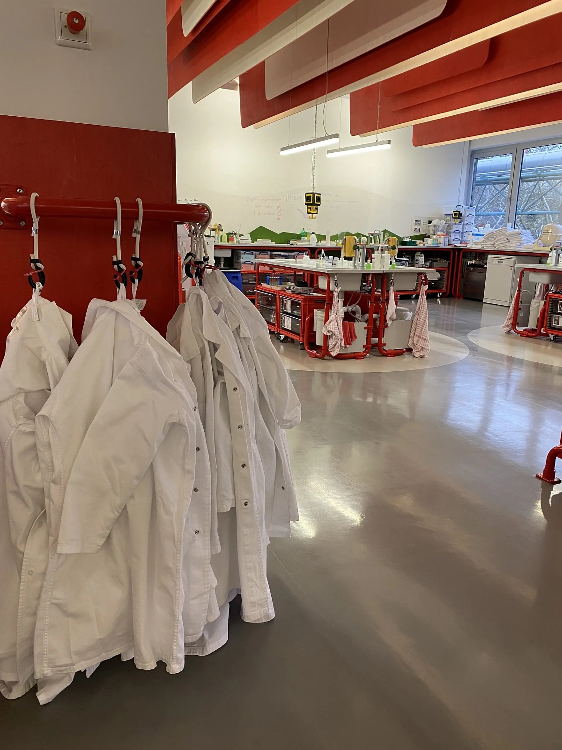 Ein moderner, heller Laborraum mit Fokus auf weißen Laborkitteln, die im Vordergrund an einer Wandhalterung hängen. Im Hintergrund befinden sich mobile Experimentierstationen mit Rohrgestellen, Arbeitsplatten und integrierten Waschbecken.