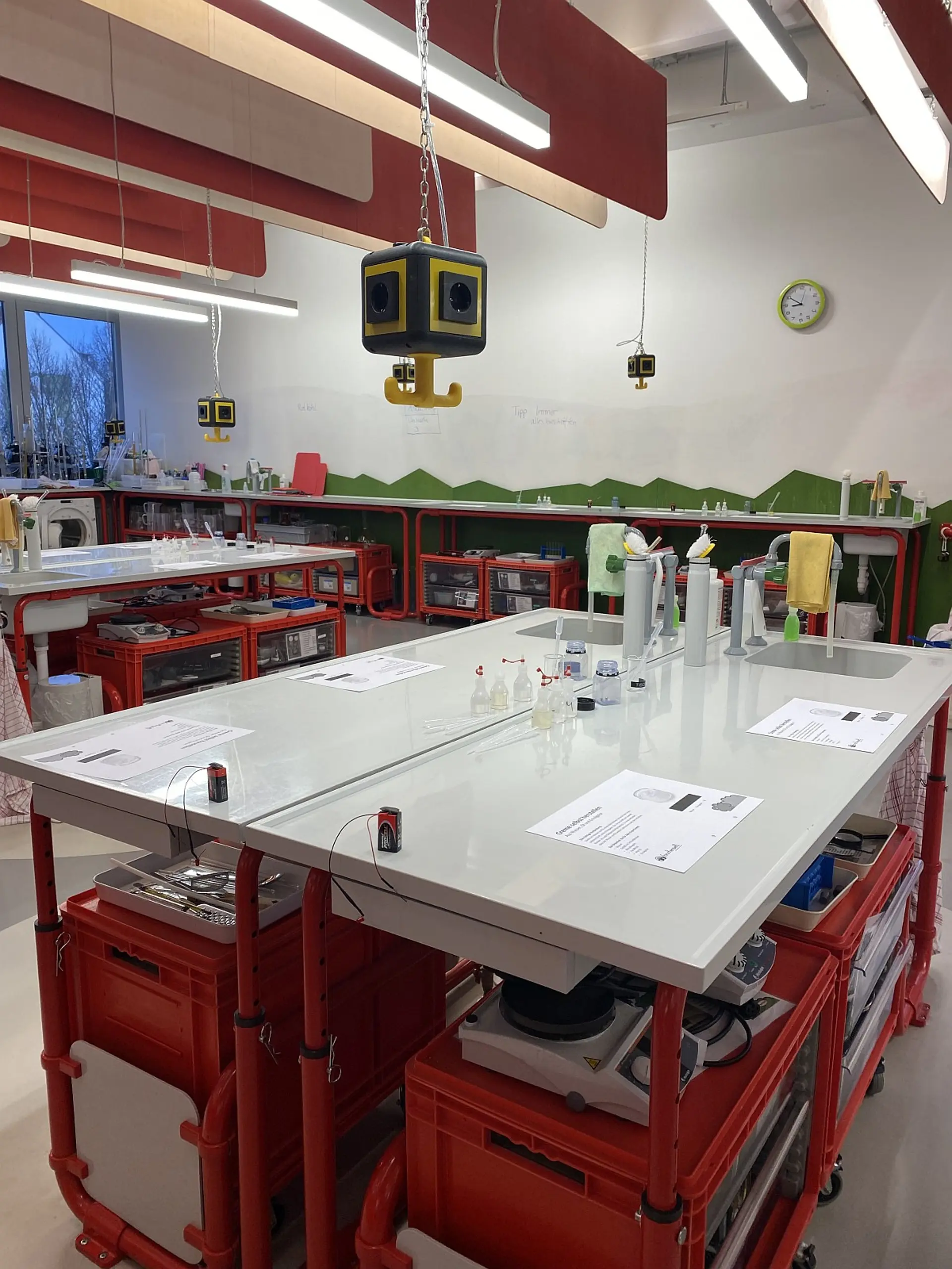 Weitwinkelaufnahme eines modernen Schullabors mit mehreren Experimentiertischen auf Rollgestellen. Jeder Tisch ist mit Arbeitsblättern, Pipetten, kleinen Bechergläsern und Batterien für Versuche vorbereitet. Von der Decke hängen Stromwürfel herab.