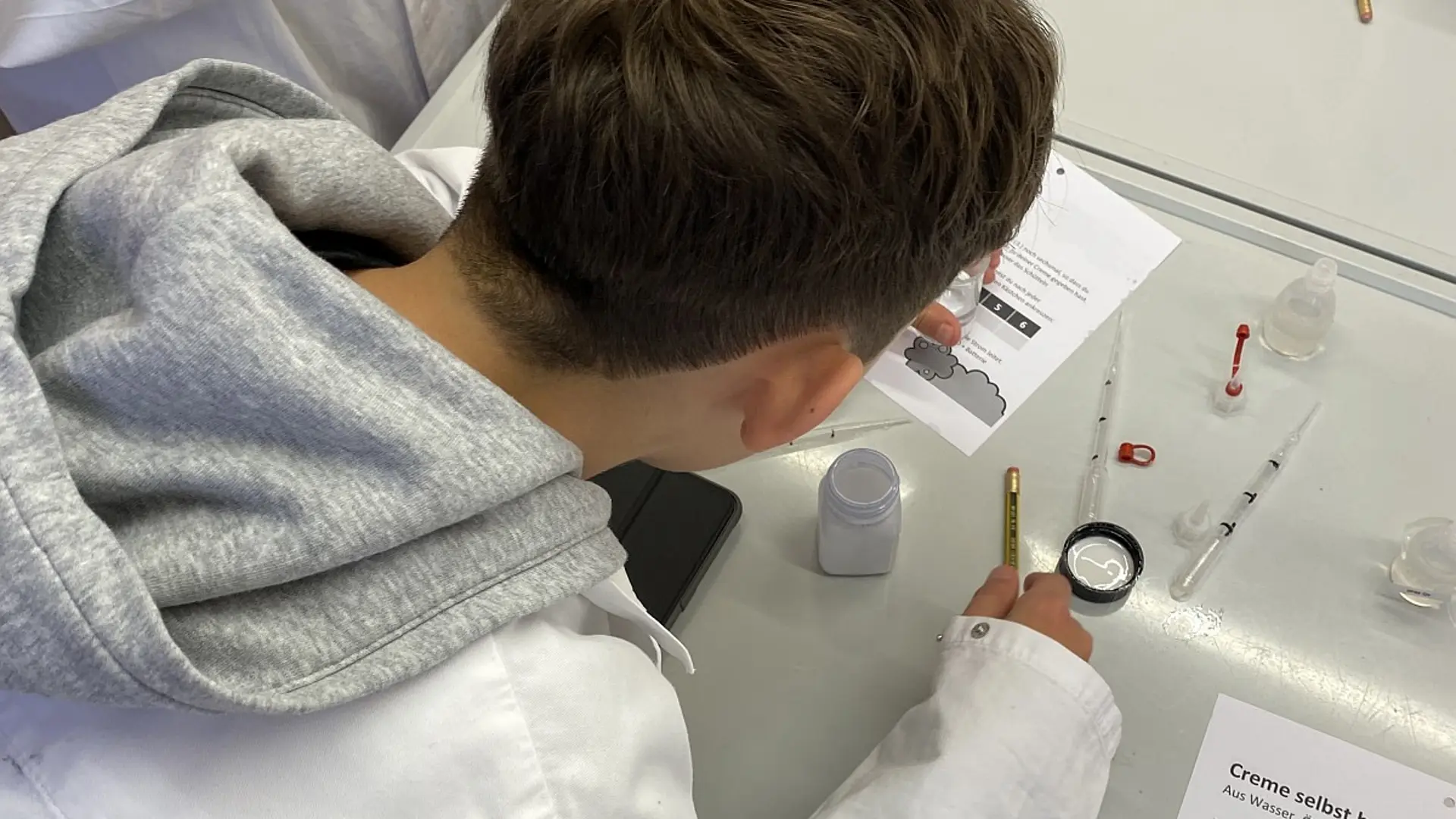 Nahaufnahme von oben eines Schülers im Laborkittel, der konzentriert an einem Experiment arbeitet. Auf dem Labortisch liegen eine detaillierte Anleitung mit dem Titel „Creme selbst herstellen“, Pipetten, kleine Behälter und ein Stift.