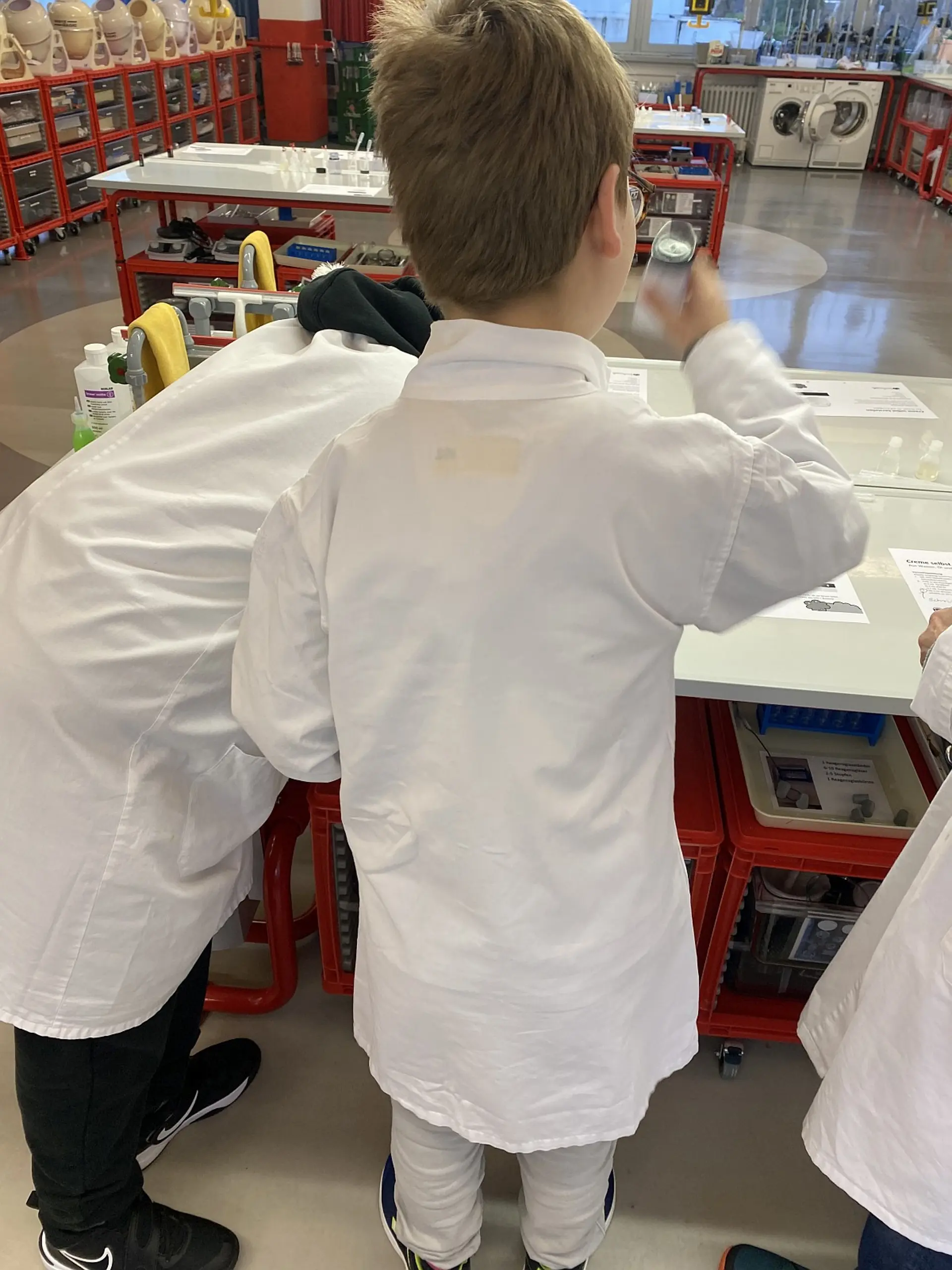 Drei Kinder in Laborkitteln stehen an einem Experimentiertisch in einem modern ausgestatteten Forscherlabor. Ein Kind im Vordergrund hält eine Messflasche vor das Gesicht, während ein anderes Kind sich konzentriert über die Arbeitsfläche beugt.