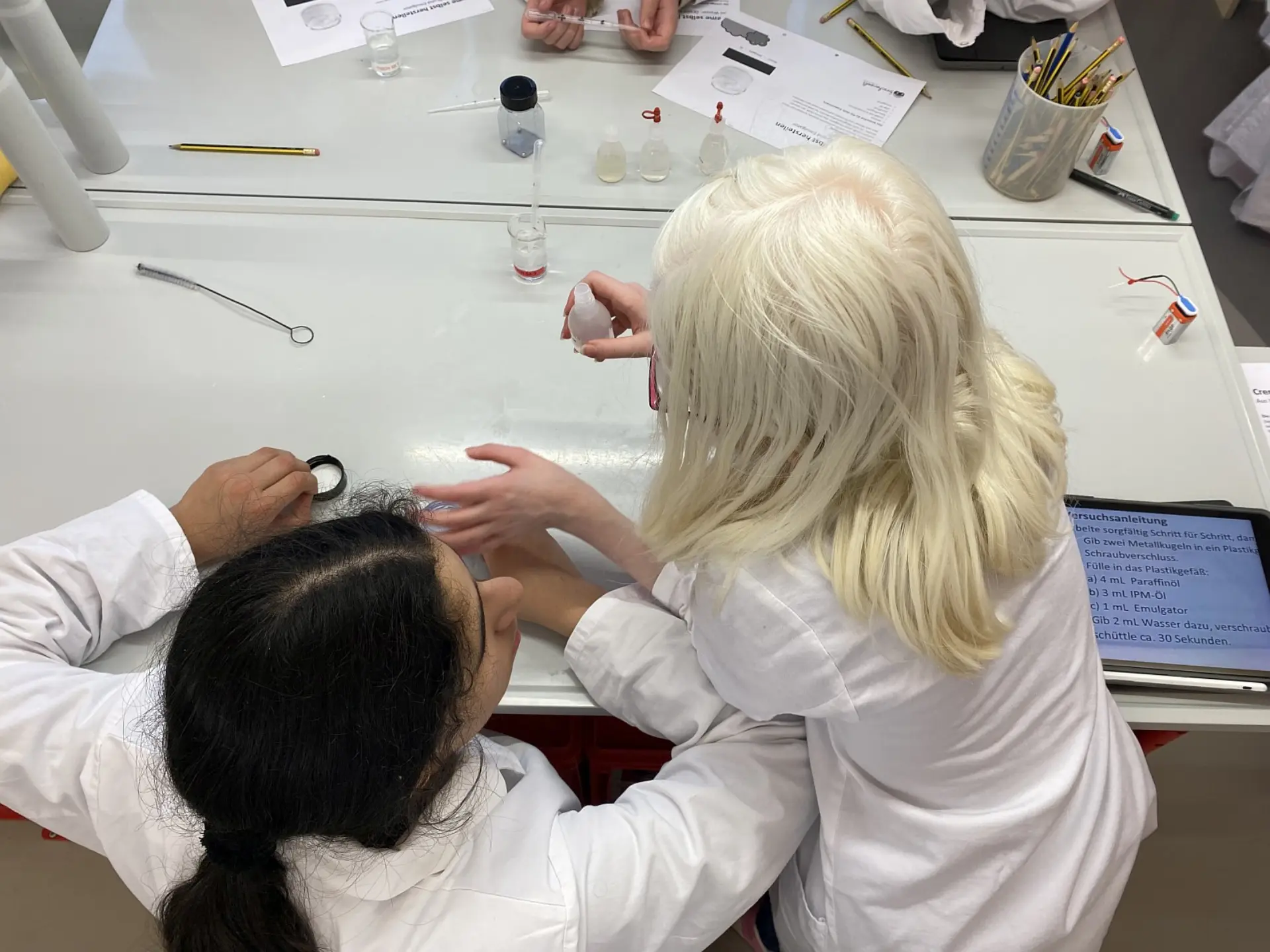 Draufsicht auf zwei Schülerinnen in Laborkitteln, die gemeinsam an einem Experimentiertisch arbeiten. Eine Schülerin hält ein kleines Gefäß, während die andere zuschaut. Auf dem Tisch liegen Pipetten, kleine Fläschchen mit Flüssigkeiten, Bleistifte und eine gedruckte Anleitung. Ein Tablet am Rand zeigt ebenfalls die Versuchsdaten an.