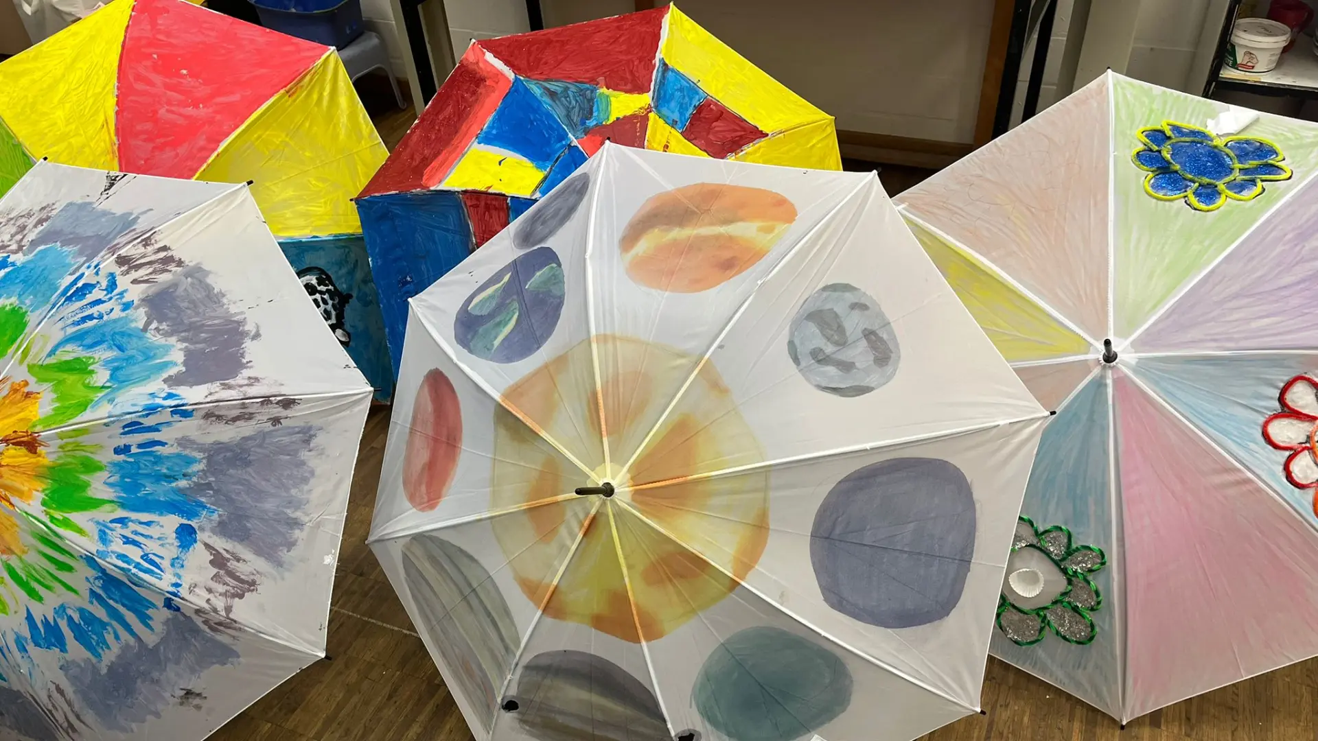 Fünf aufgespannte Regenschirme liegen auf einem Holzboden und zeigen verschiedene kreative Designs aus dem Kunstunterricht.