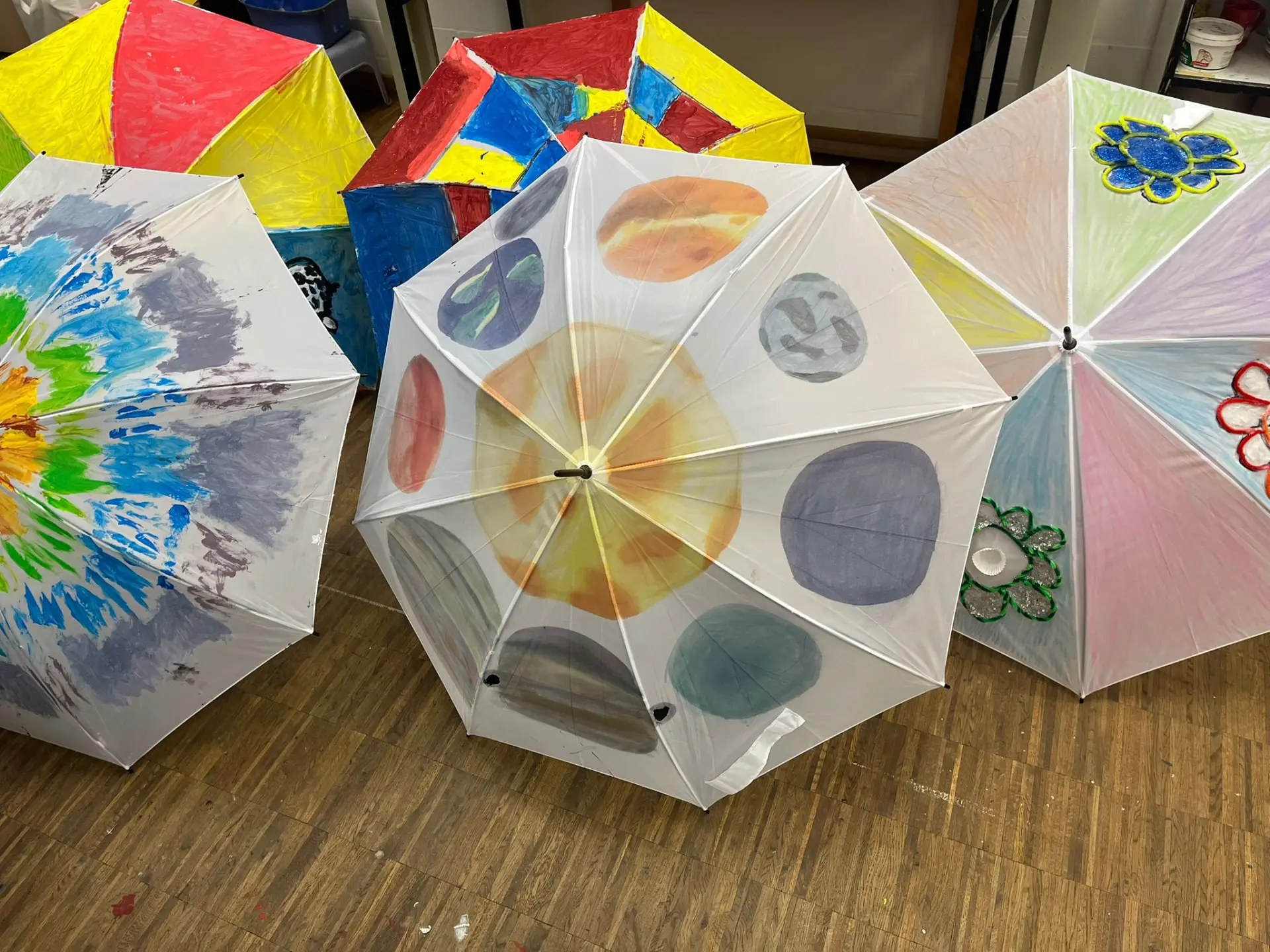 Fünf aufgespannte Regenschirme liegen auf einem Holzboden und zeigen verschiedene kreative Designs aus dem Kunstunterricht.