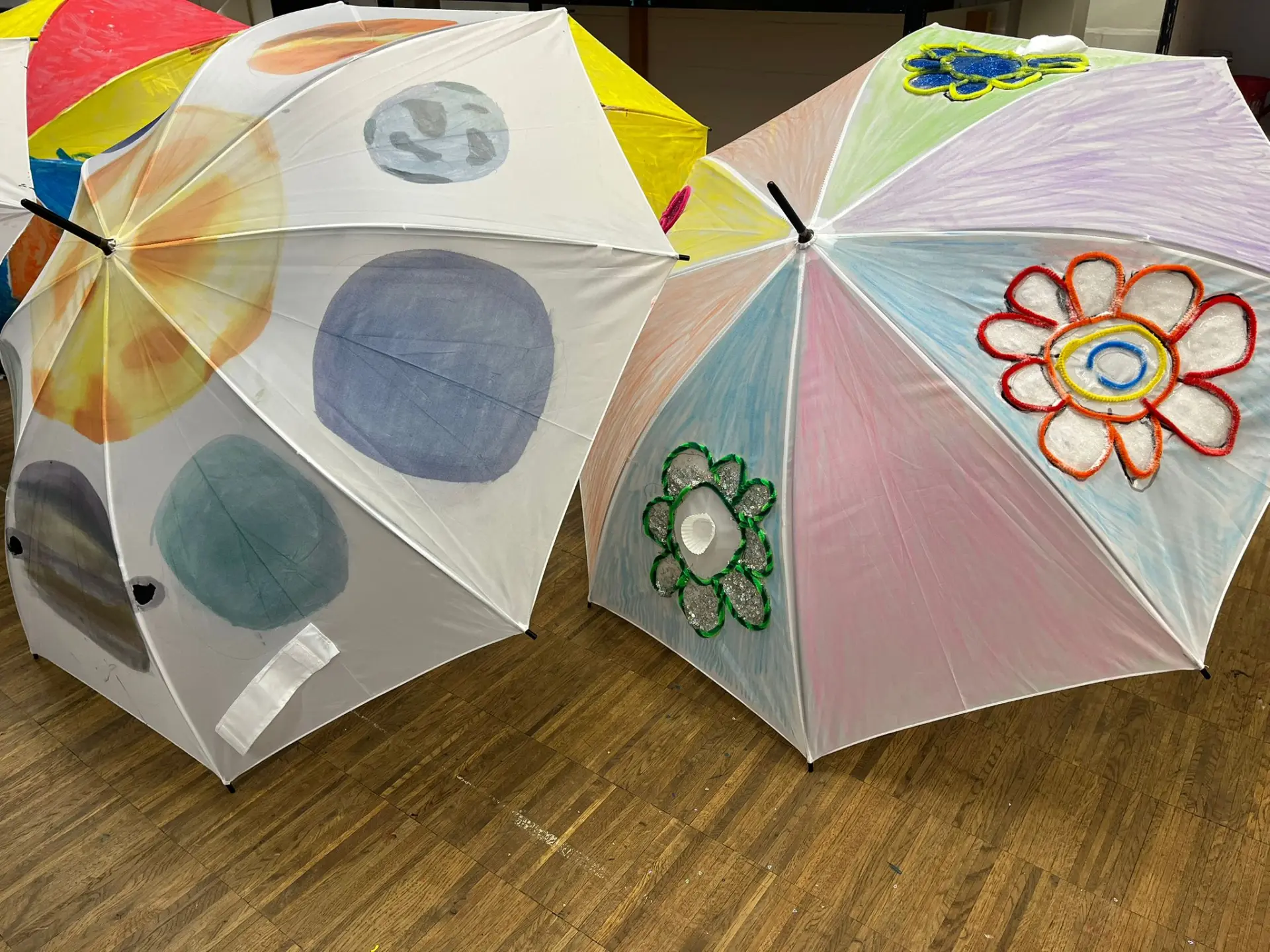 Zwei aufgespannte Regenschirme liegen auf dem Boden. Der Schirm links ist mit Planeten des Sonnensystems bemalt, der andere zeigt bunte Blumenmotive.