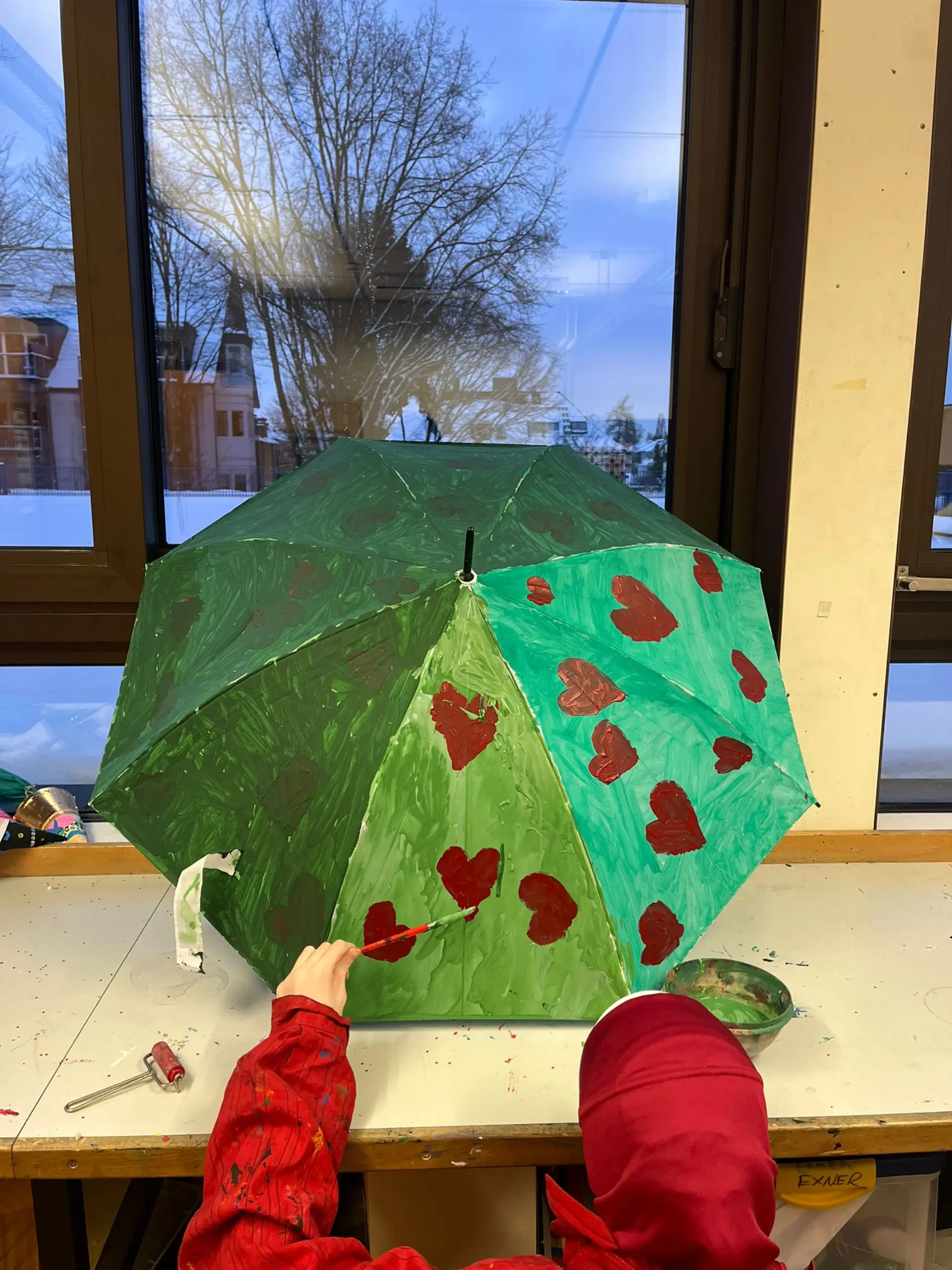 Rückansicht einer Schülerin in einem Malkittel, die in einem Kunstraum an einem Regenschirm arbeitet. Der Schirm ist in verschiedenen Grüntönen bemalt und mit roten Herzen mit einem Malpinsel verziert.