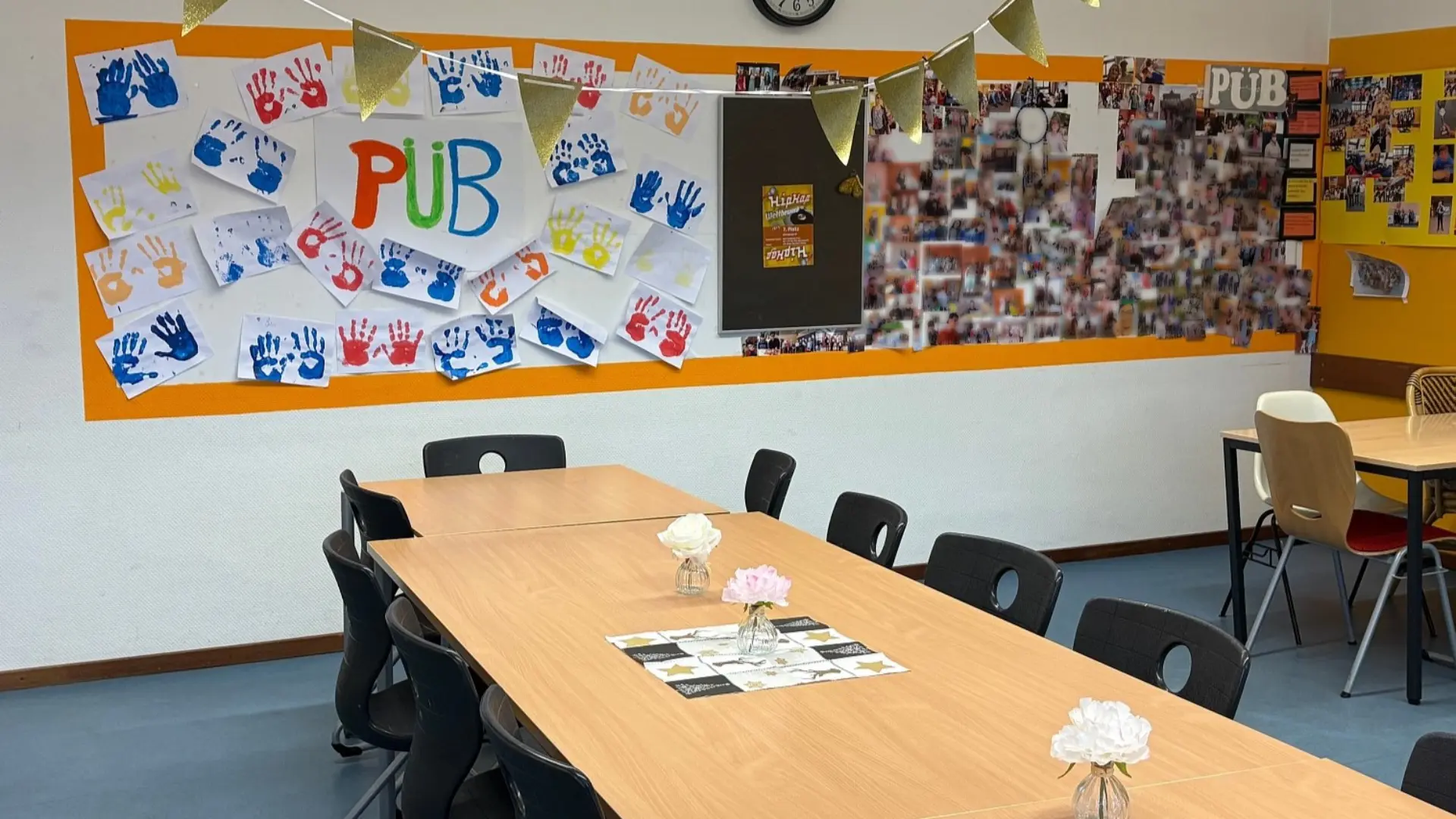Ein heller Gruppenraum mit einem langen Tisch in der Mitte, umgeben von vielen Stühlen. An der Wand im Hintergrund ist ein großes Plakat mit dem Schriftzug "PÜB" in bunten Buchstaben angebracht, umrahmt von zahlreichen bunten Handabdrücken. Rechts daneben befindet sich eine Pinnwand mit vielen Fotos und Bildern.