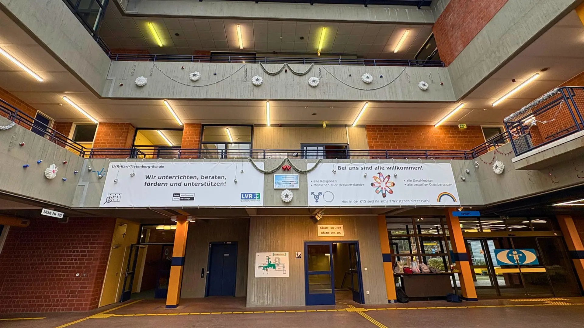 Die zweistöckige, weitläufige Eingangshalle der Schule, die bis zum verglasten Dach reicht. Das Gebäude hat eine offene Bauweise mit hellen Betonbrüstungen und Metallgeländern. Mehrere Banner hängen von der ersten Etage herab, darunter ein großes Banner mit dem Schullogo und den Slogans: 'Wir unterrichten, beraten, fördern und unterstützen!' und 'Bei uns sind alle willkommen!' mit Piktogrammen für verschiedene Gruppen.