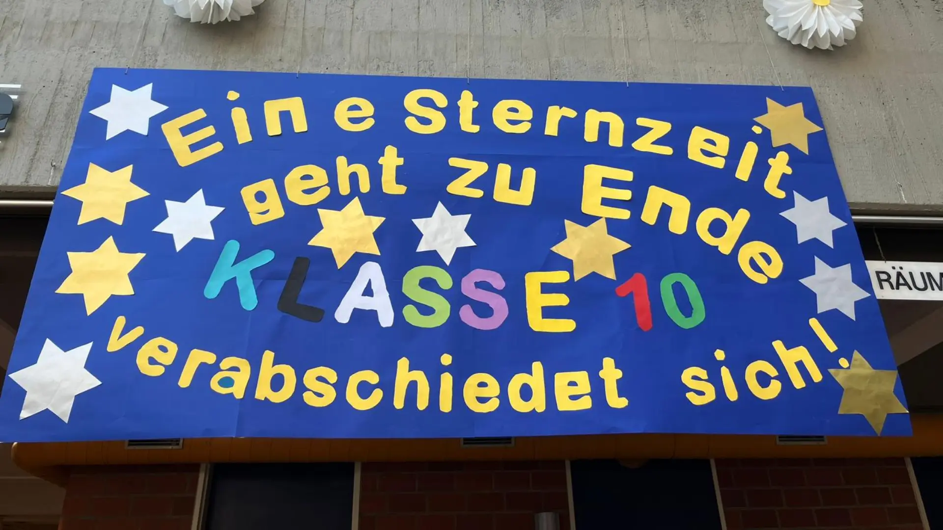 Ein großes Banner hängt an einer Betonwand im Eingangsbereich der Schule. Darauf steht in bunten Buchstaben: Eine Sternzeit geht zu Ende – Klasse 10 verabschiedet sich! Das Banner ist mit zahlreichen Sternen verziert.