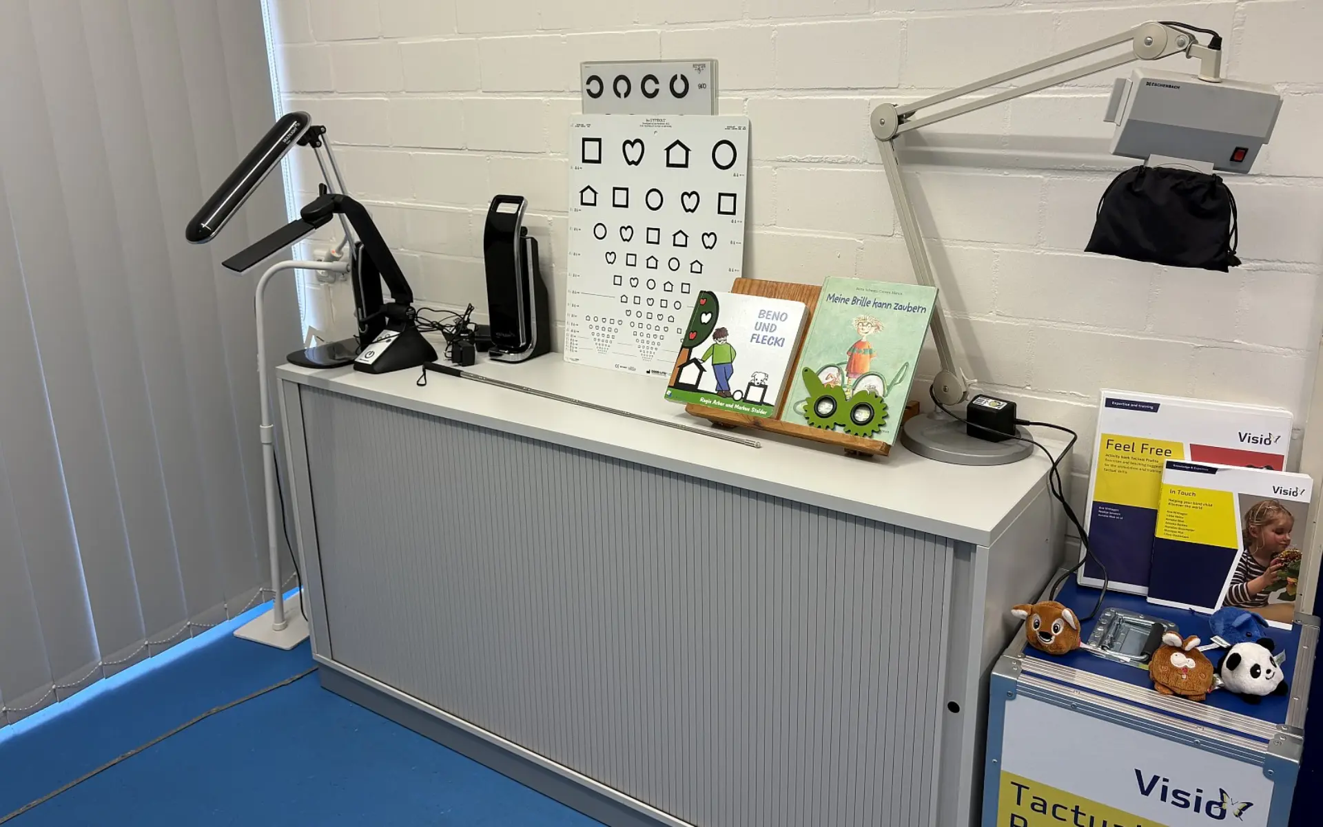 Ein Sideboard, auf dem verschiedene Materialien für die Sehberatung angeordnet sind. Zu sehen sind Sehzeichen-Tafeln mit Symbolen, Kinderbücher zum Thema Sehen und Brillen sowie Blindenstock.