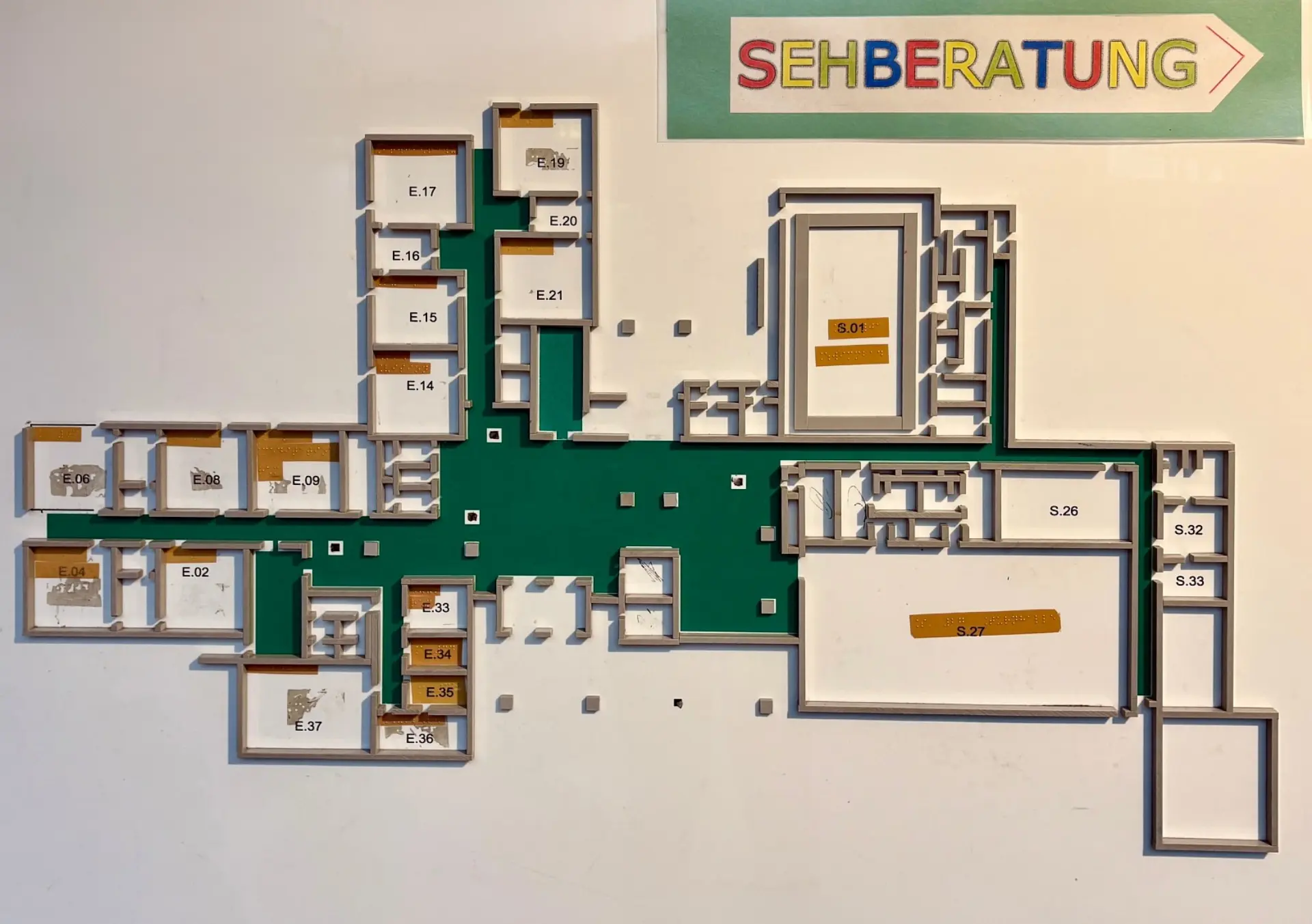 Ein taktiler Lageplan der LVR-Karl-Tietenberg-Schule, der an einer Wand hängt. Der Plan zeigt den Grundriss des Schulgebäudes als dreidimensionale Darstellung. Oben rechts weist ein Schild mit der Aufschrift 'Sehberatung' in bunten Buchstaben und einem Pfeil nach rechts auf den Standort der Sehberatung hin.