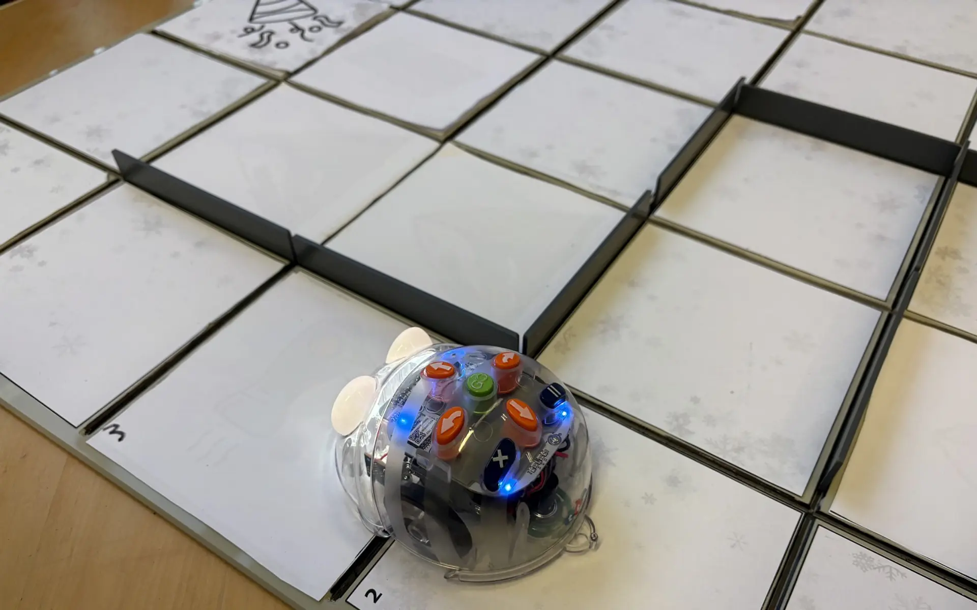 Ein kleiner, transparenter Lernroboter (Blue-Bot) mit orangefarbenen Tasten und blau leuchtenden Lichtern auf einem Spielfeld. Das Spielfeld besteht aus einem Raster mit quadratischen Feldern, auf denen teilweise Schneeflocken-Muster zu sehen sind.
