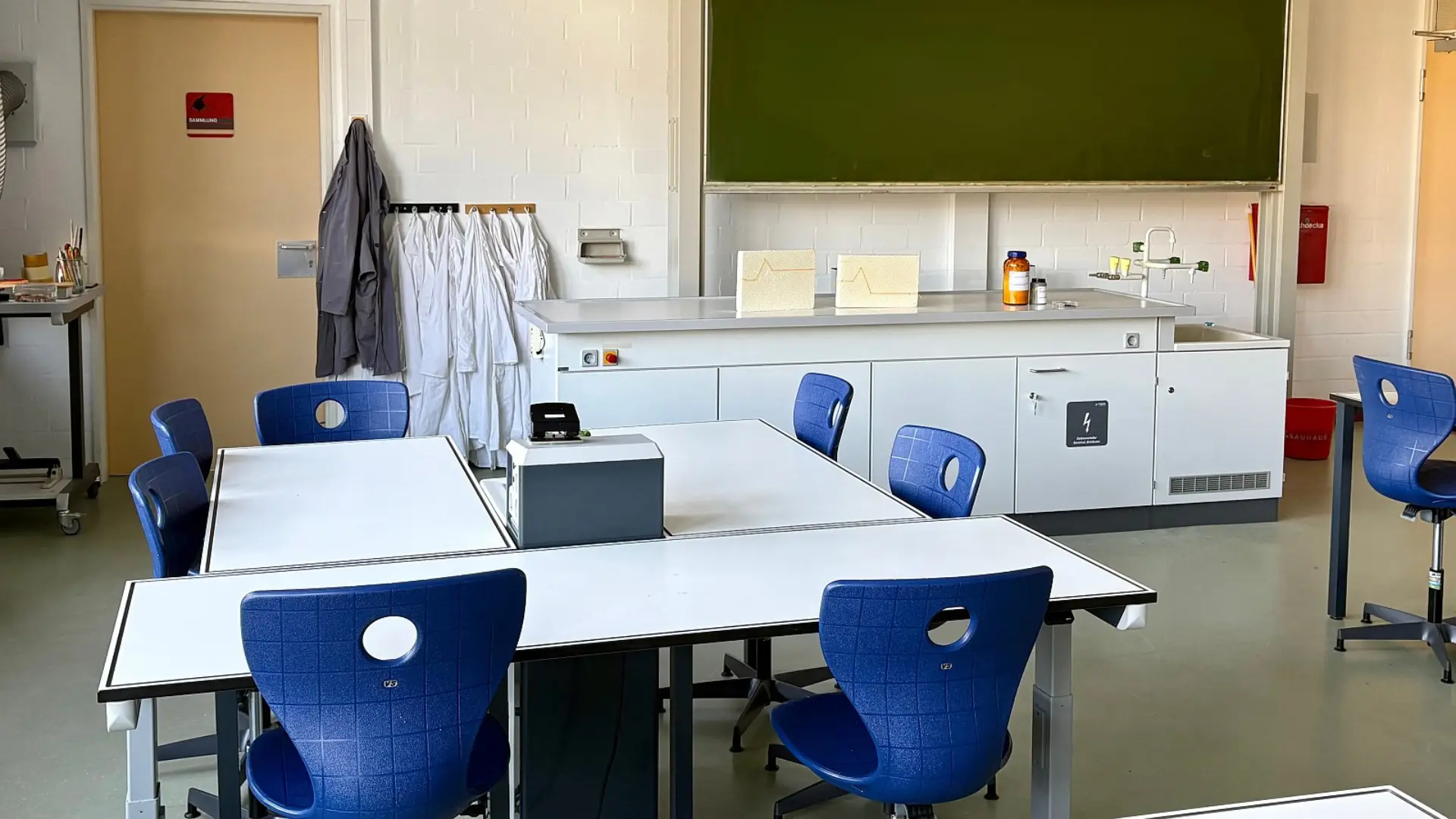 Ein heller Naturwissenschaftsraum mit einem großen Lehrerpult vor einer Schreibtafel. Auf dem Pult stehen Versuchsaufbauten und Chemikalienflaschen. Im Vordergrund gruppieren sich Schülertische mit Stühlen. An der Ziegelwand hängen mehrere Laborkittel bereit.