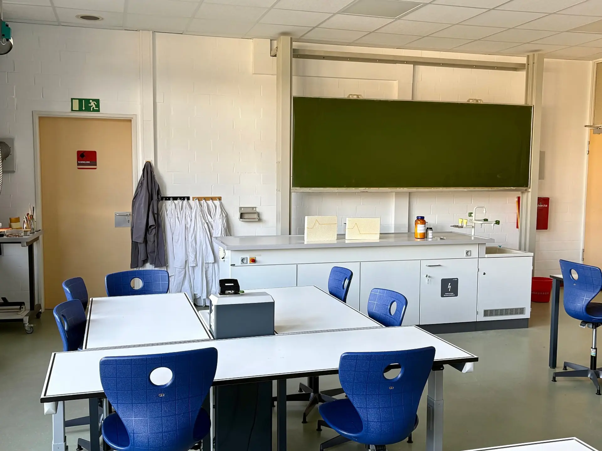 Ein heller Naturwissenschaftsraum mit einem großen Lehrerpult vor einer Schreibtafel. Auf dem Pult stehen Versuchsaufbauten und Chemikalienflaschen. Im Vordergrund gruppieren sich Schülertische mit Stühlen. An der Ziegelwand hängen mehrere Laborkittel bereit.