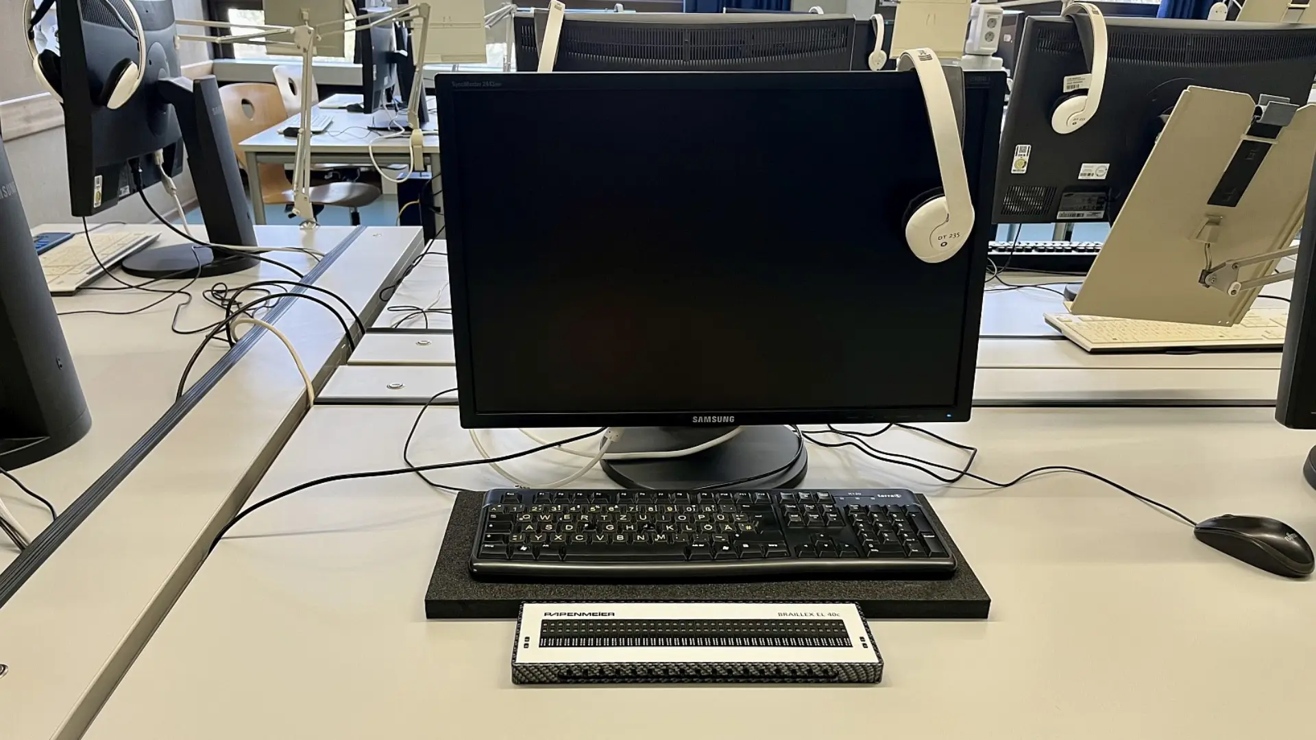 Ein barrierefreier Computerarbeitsplatz in einem PC-Raum. Vor einem Monitor liegt eine Tastatur und davor eine Braillezeile. Am Monitor hängt ein Kopfhörer. Im Hintergrund sind weitere ähnliche Arbeitsstationen mit Monitoren, Halterungen für Tablets und Kopfhörern zu sehen.