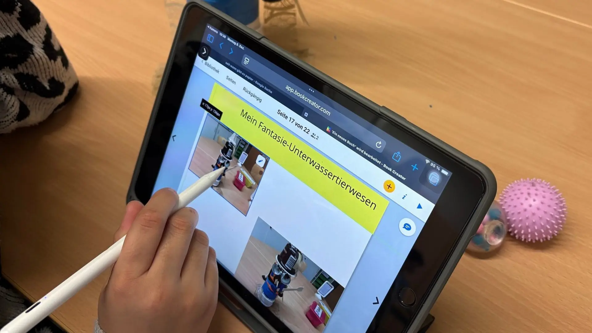 Nahaufnahme der Hand eines Kindes der Primarstufe, das ein iPad auf einem Holztisch bedient. Das Kind hält einen Eingabestift und interagiert mit der App 'Book Creator'. Auf dem Bildschirm ist die Seite 17 von 22 eines digitalen Buches mit dem Titel 'Mein Fantasie-Unterwassertierwesen' zu sehen.