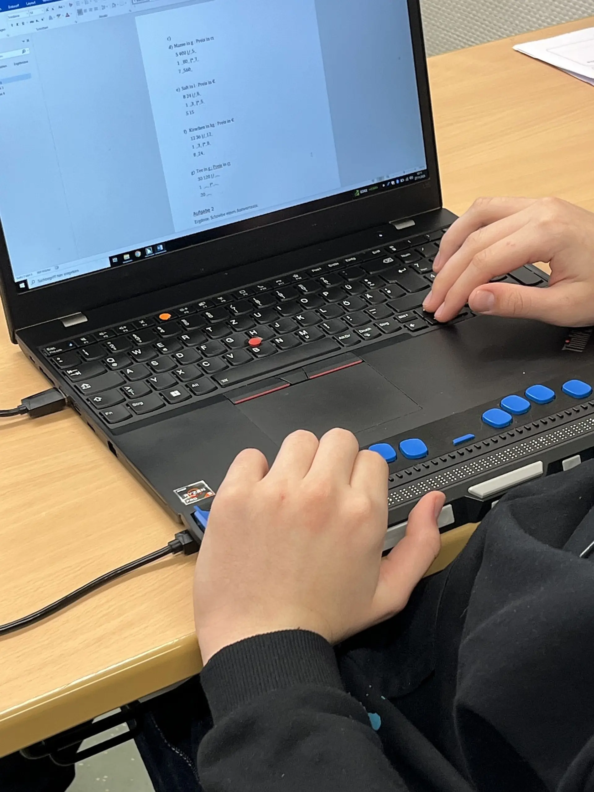 Nahaufnahme eines Laptops auf einem Holztisch, der von einem Schüler oder einer Schülerin der Sekundarstufe genutzt wird. Am unteren Rand des Laptops ist ein externes Braille-Zeilendisplay angeschlossen. Auf dem Laptop-Bildschirm sind Matheaufgaben zu sehen. 