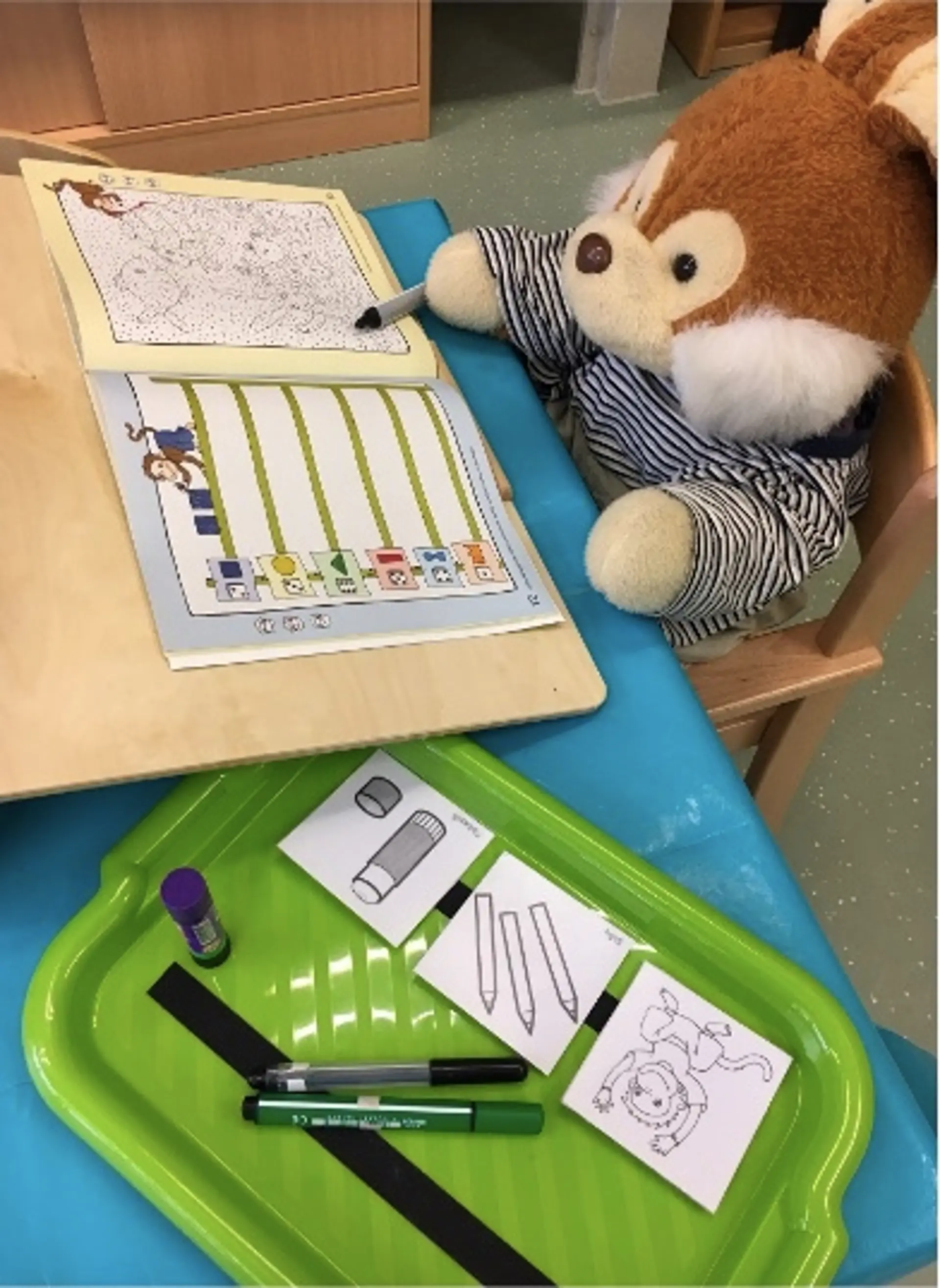 Ein Lernarbeitsplatz in der Vorschule mit der Stoffpuppe Harry Hase, die an einem kleinen Holztisch sitzt und ein Arbeitsheft bearbeitet.Vor ihm liegt ein Kunststofftablett mit Stiften, einem Klebestift und Bildkarten, die Schulutensilien wie Stifte und Kleber zeigen.